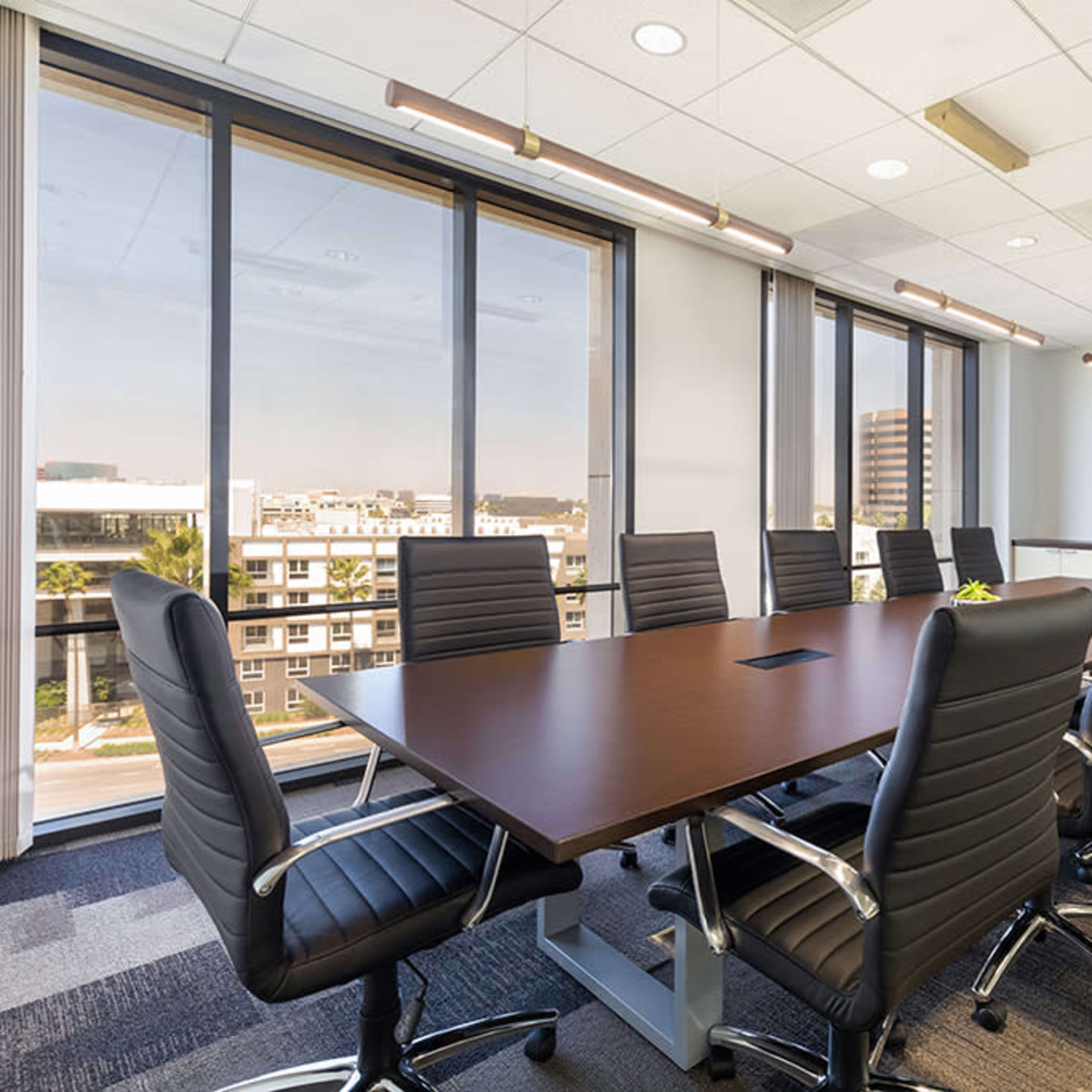 A modern conference room features a long table surrounded by black leather chairs, with large windows that provide natural light and a flat-screen TV mounted on the wall.