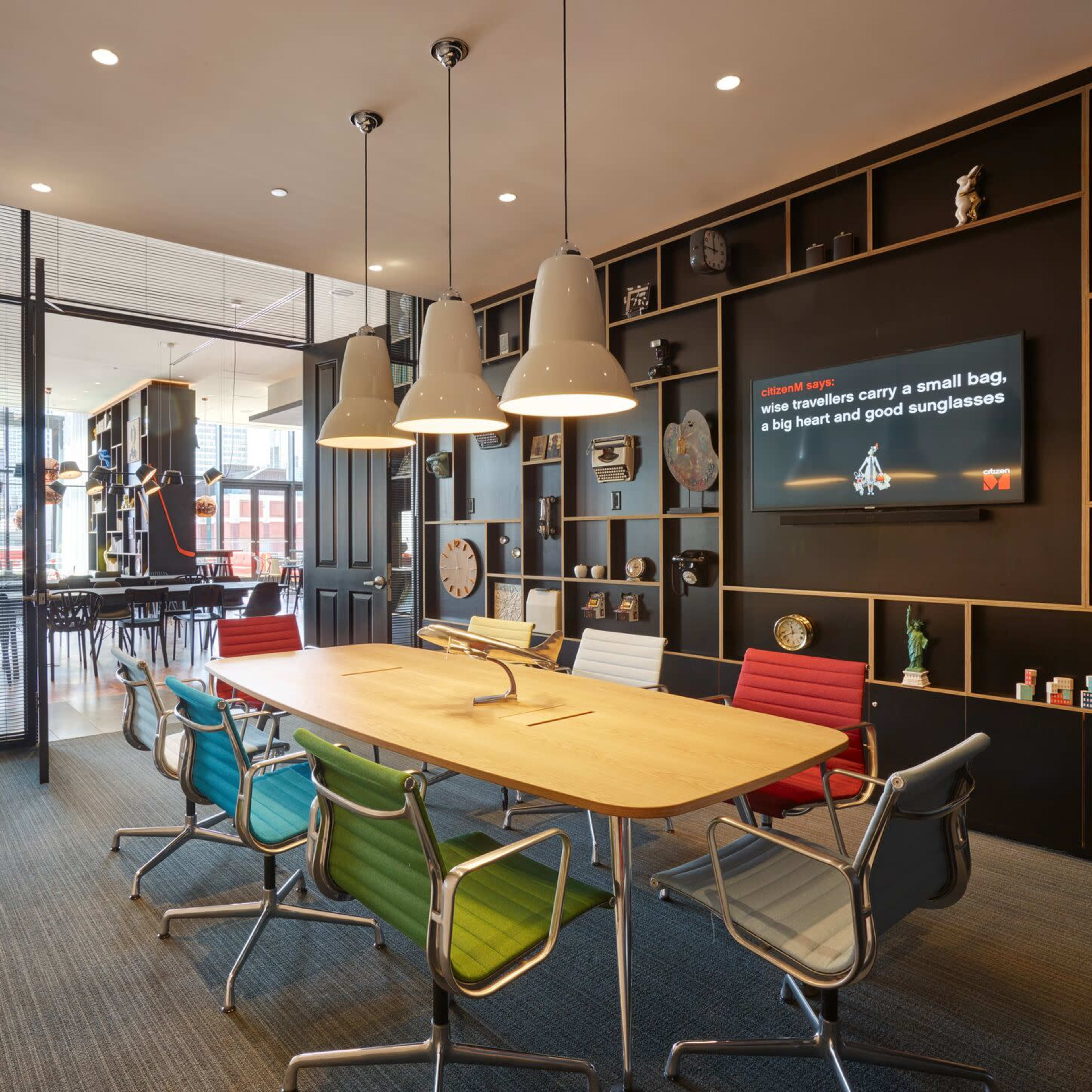 A modern conference room features a large wooden table surrounded by colorful chairs and a wall-mounted screen, with floor-to-ceiling windows providing a view of the outside.