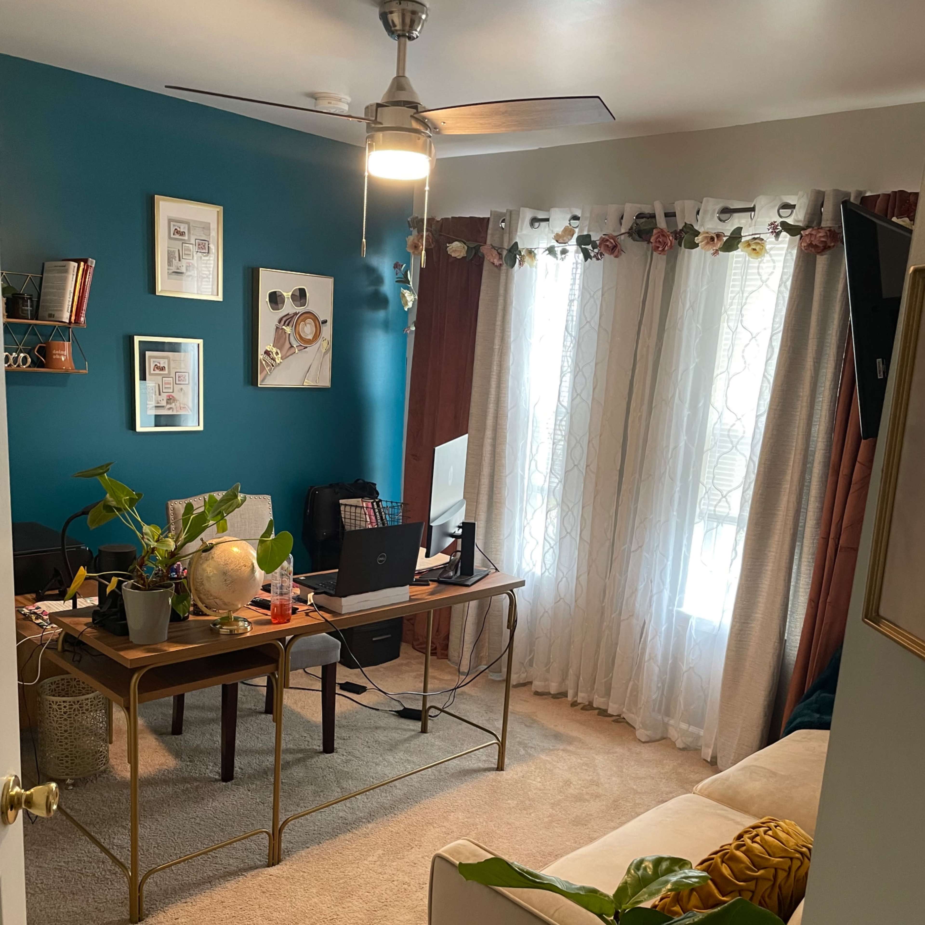 A cozy home office with a desk, computer, and various plants, set against a teal wall and bright window treatments.