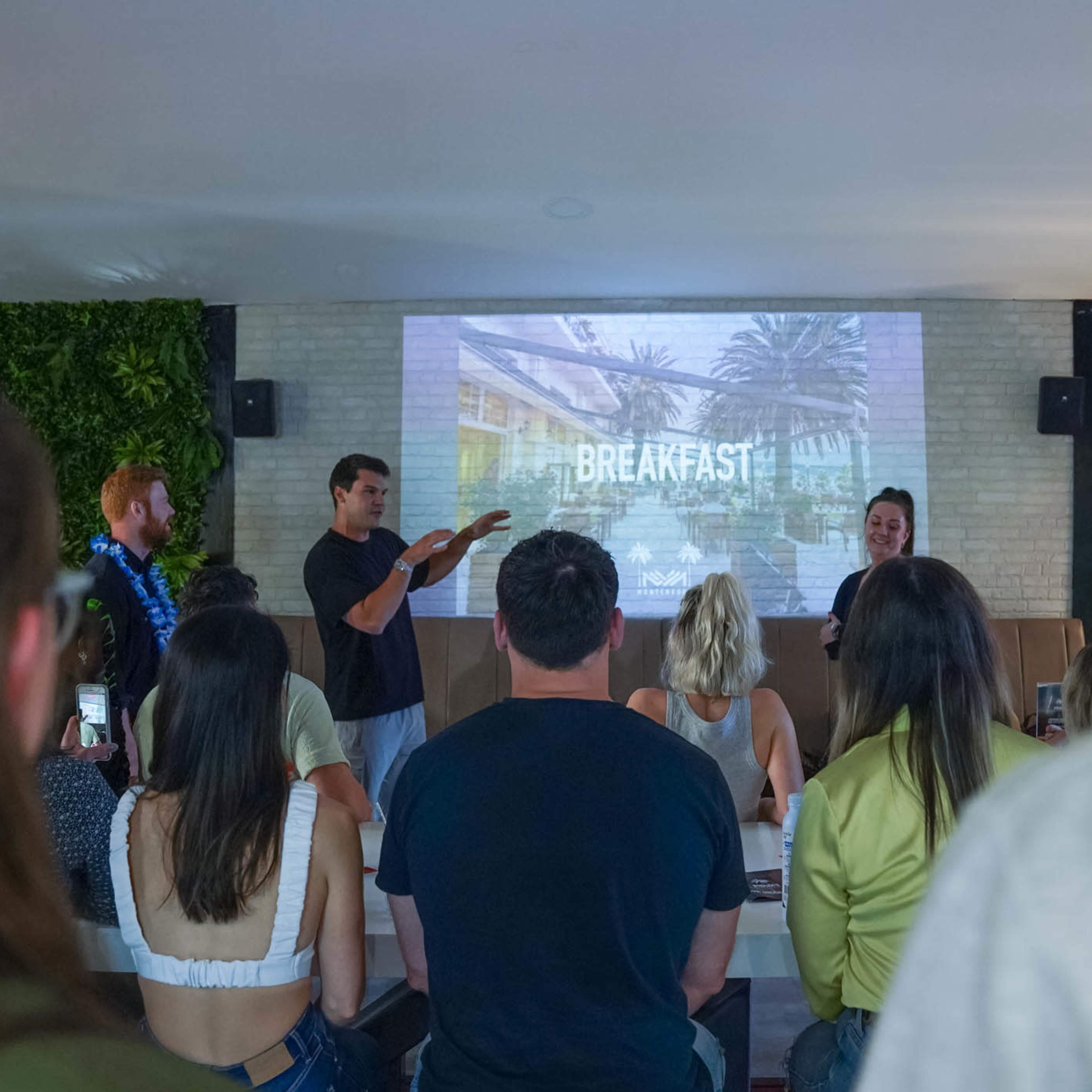 A group of people is gathered in a room for a presentation on breakfast, with one speaker gesturing towards a screen displaying information.