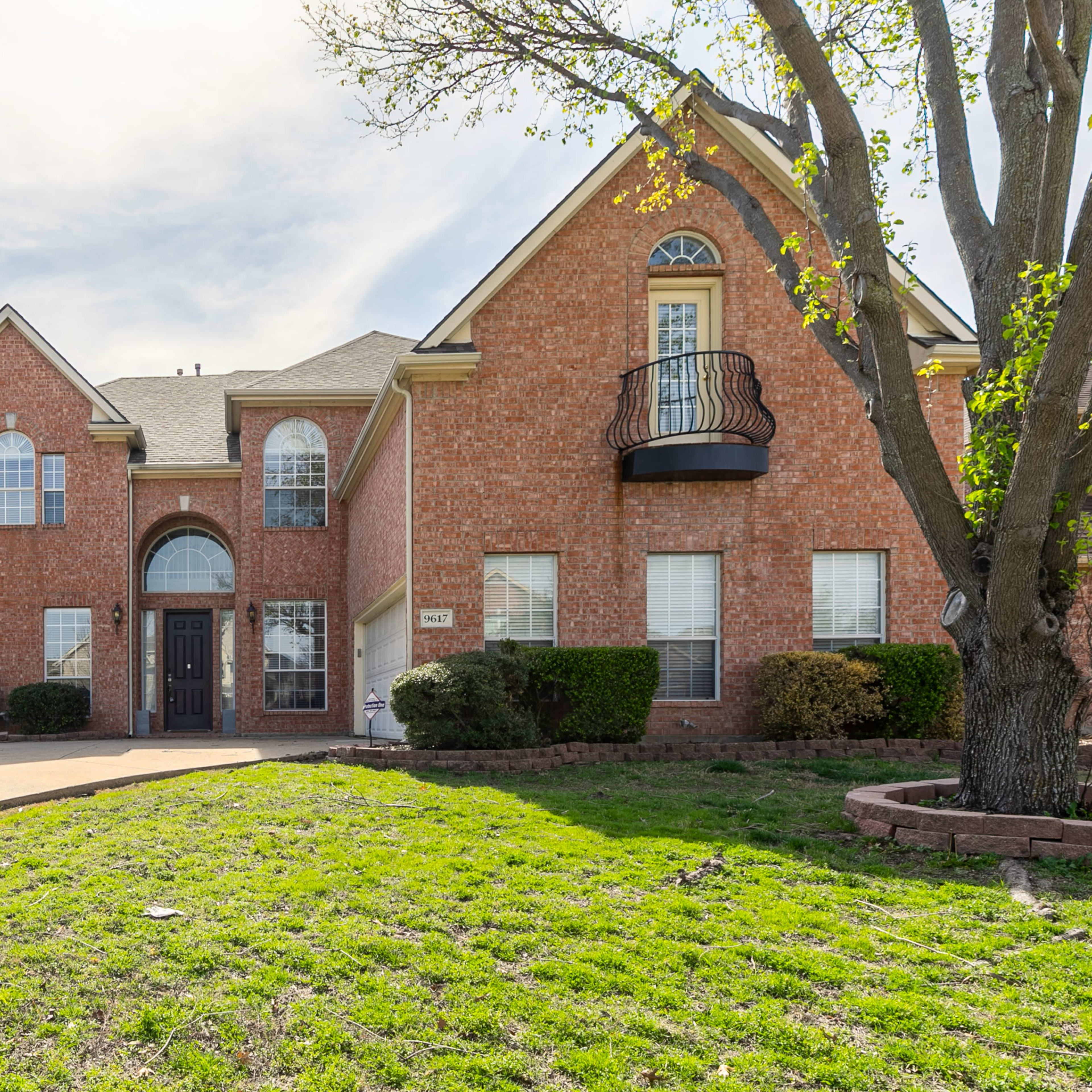 A two-story brick house with a front balcony, a well-manicured lawn, and a driveway leading to the entrance.