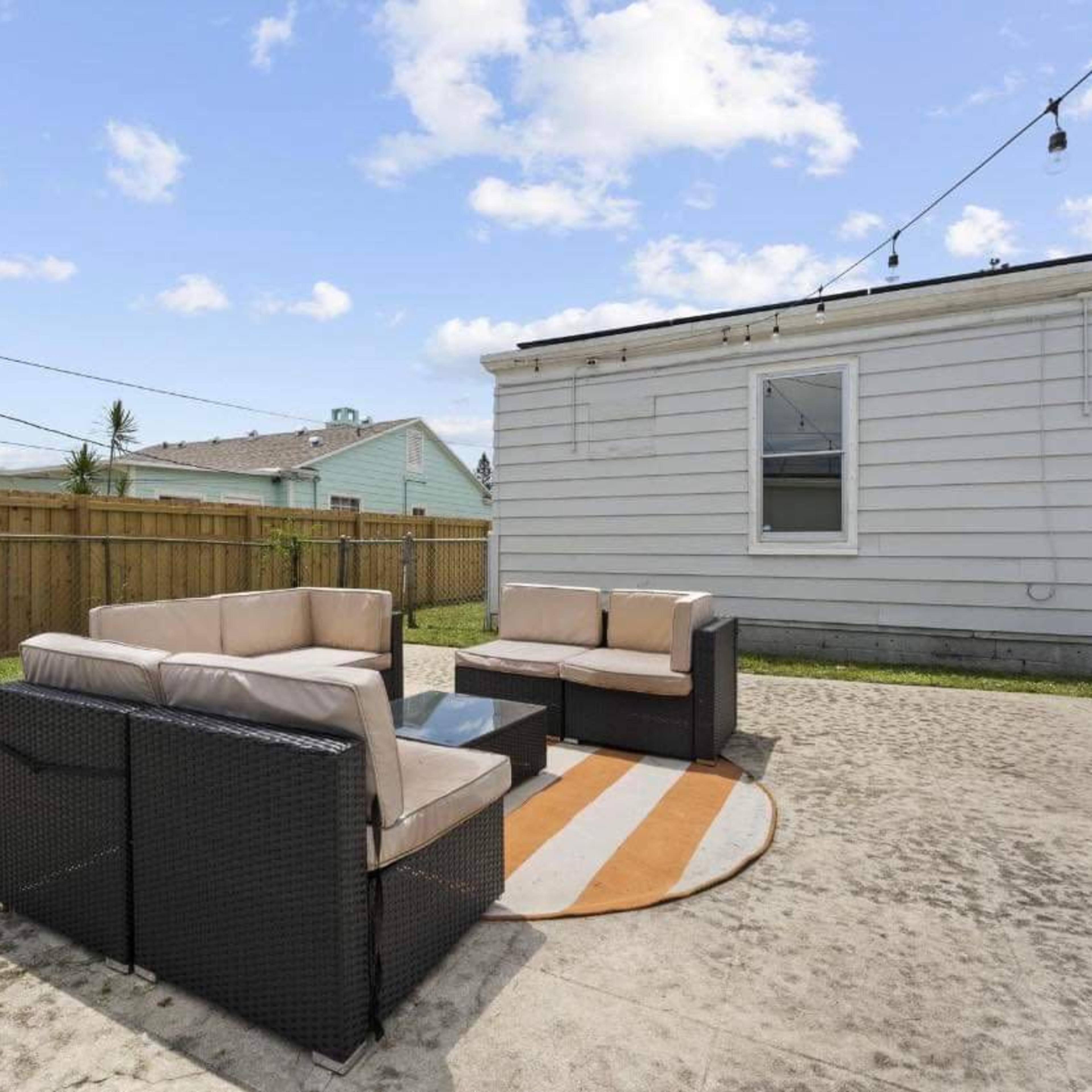 A backyard features a seating area with a sectional sofa and a coffee table on a circular rug, surrounded by grass and a wooden fence.