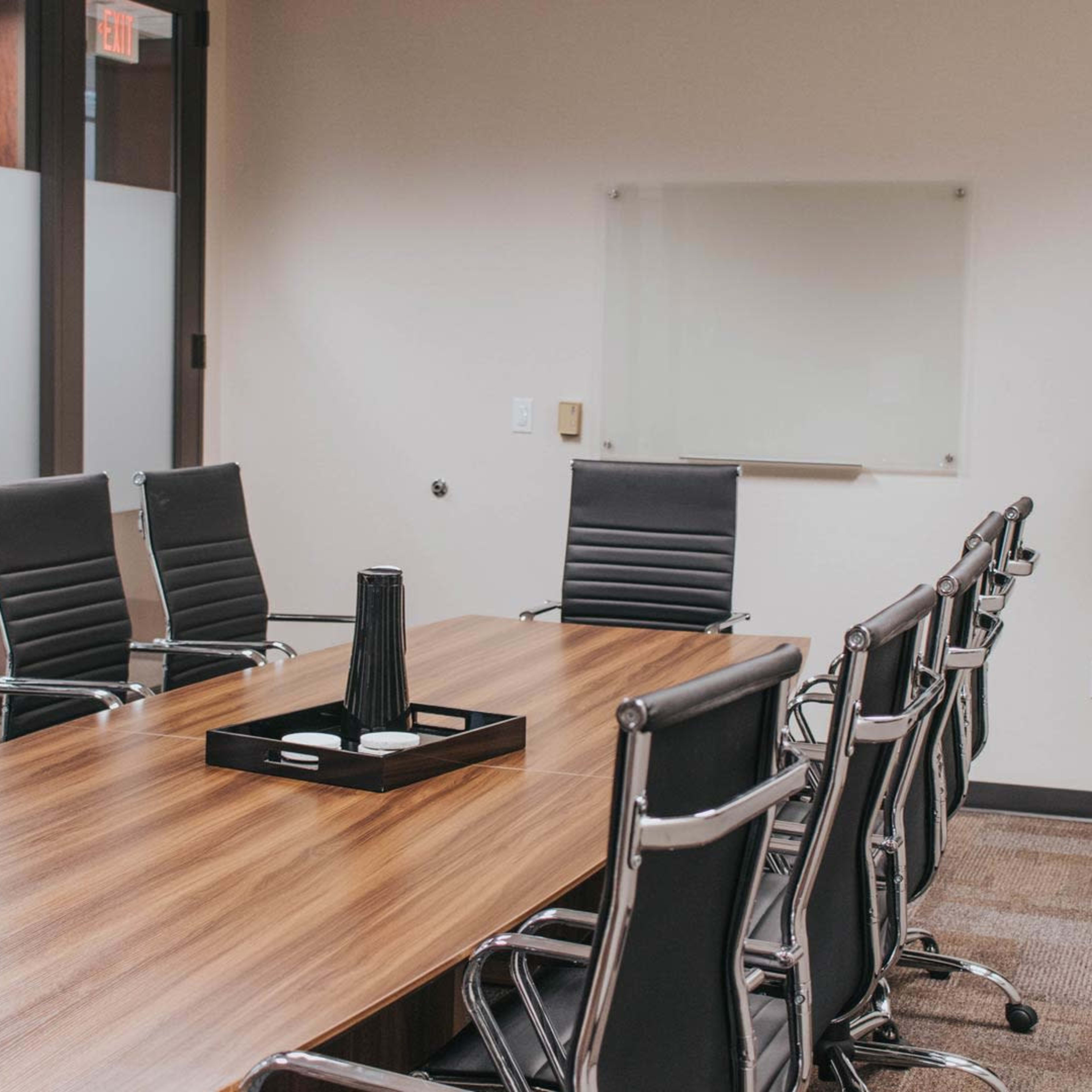 A modern conference room features a long wooden table surrounded by black ergonomic chairs, with a potted plant in one corner and a glass whiteboard on the wall.