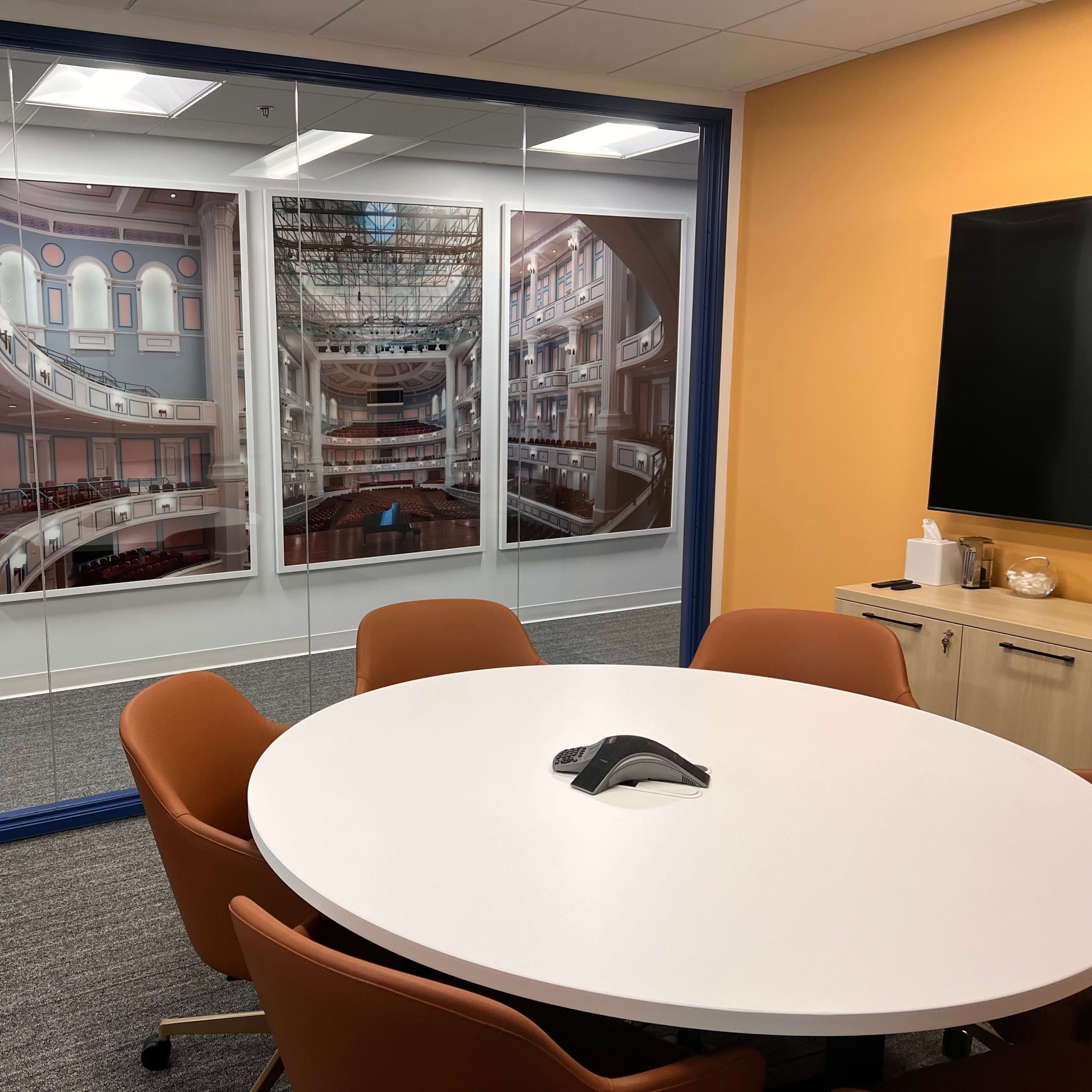 A conference room with a round white table surrounded by orange chairs and large framed photographs of a theater on the walls.