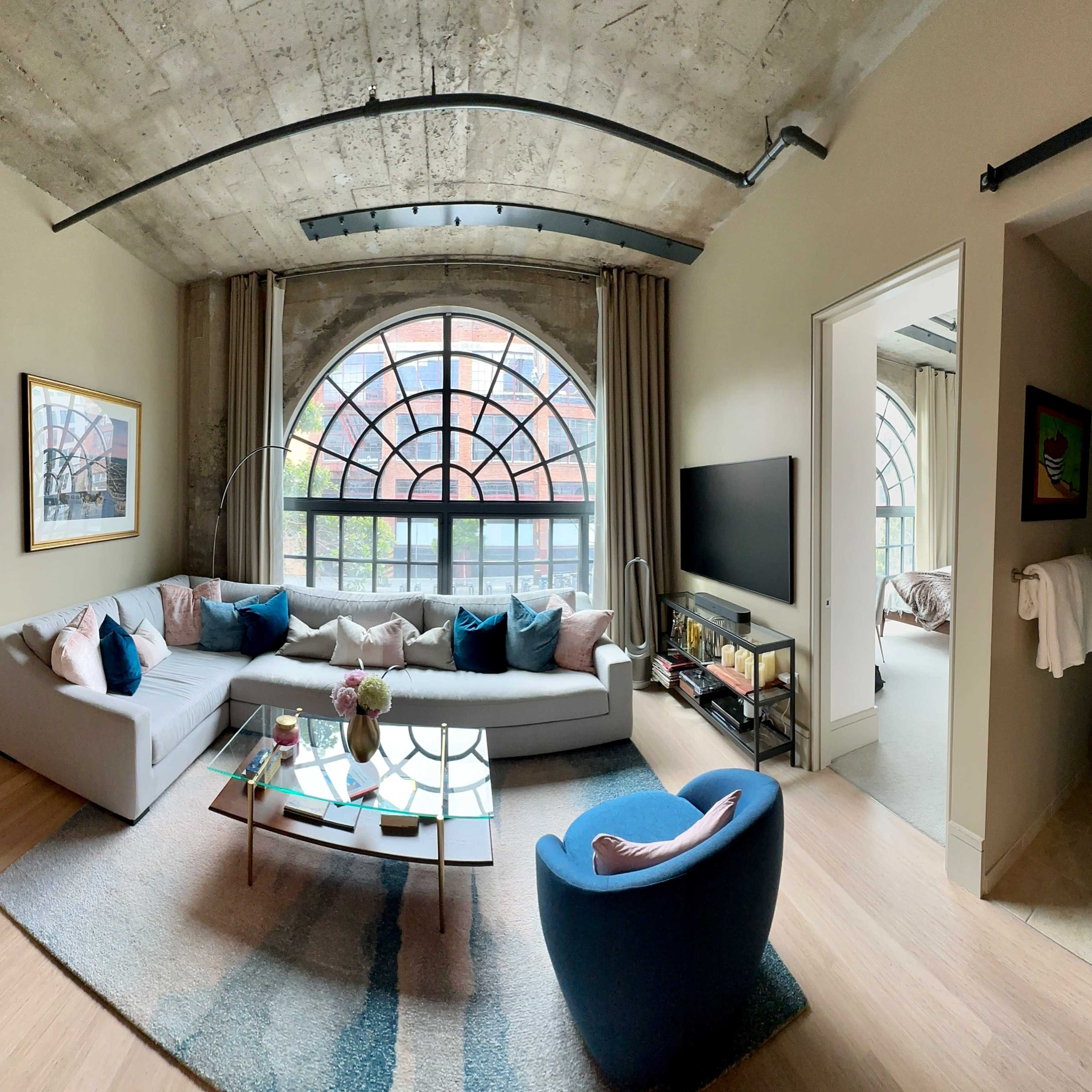 The living room features a large sectional sofa, a round coffee table, and a window with an architectural arch, providing views of the outdoors.