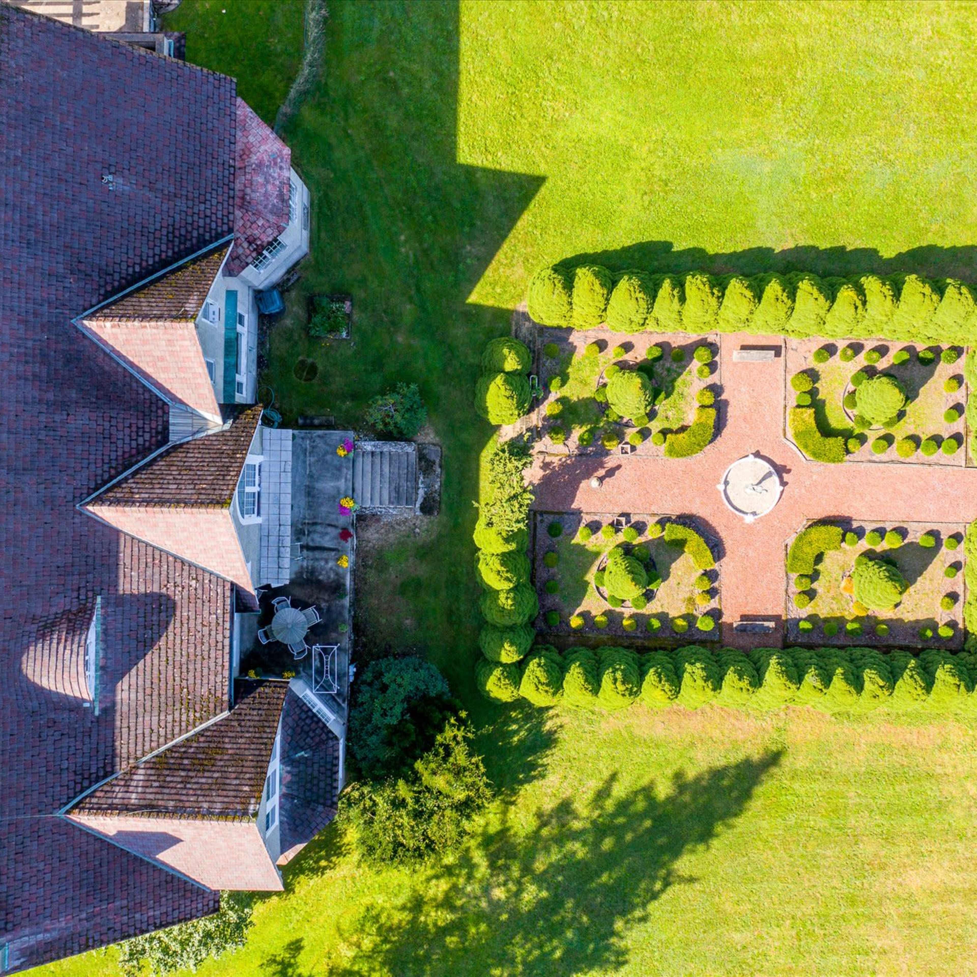 An aerial view shows a large house adjacent to a neatly landscaped garden with symmetrical trees and a central stone feature.