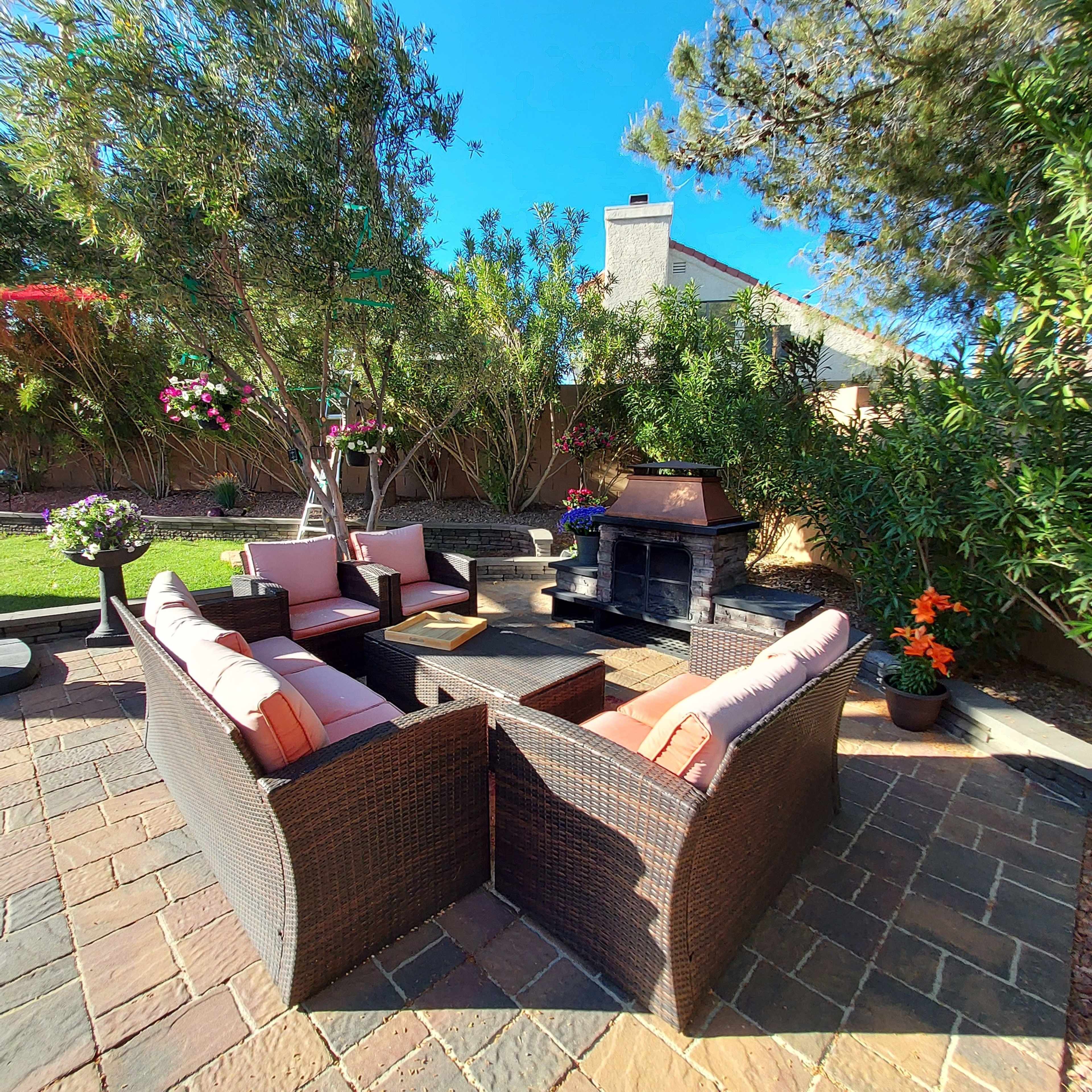 A cozy outdoor seating area features wicker furniture arranged around a stone fireplace, surrounded by plants and trees.