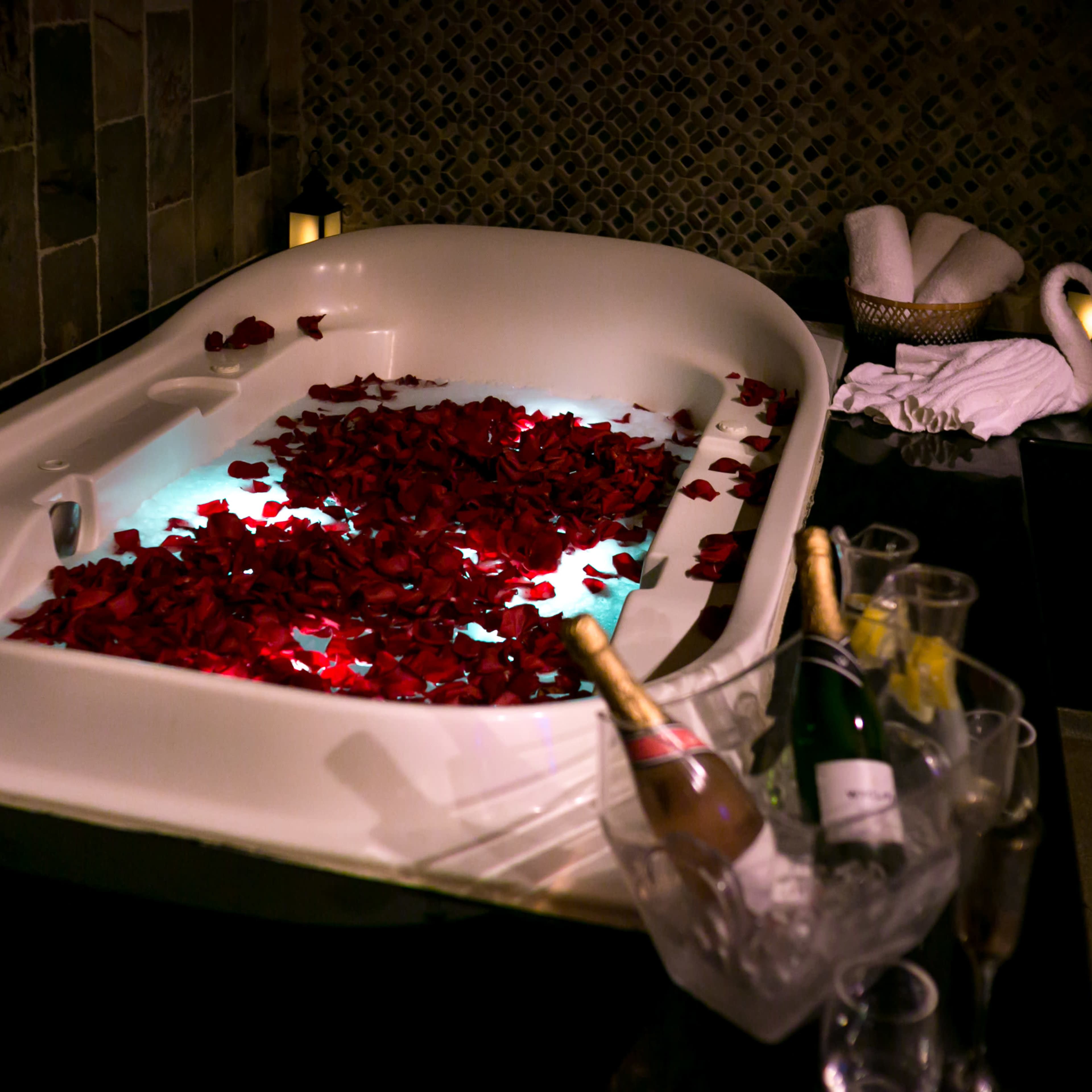 A white bathtub filled with rose petals and illuminated with soft lighting sits beside a champagne bucket and glasses.