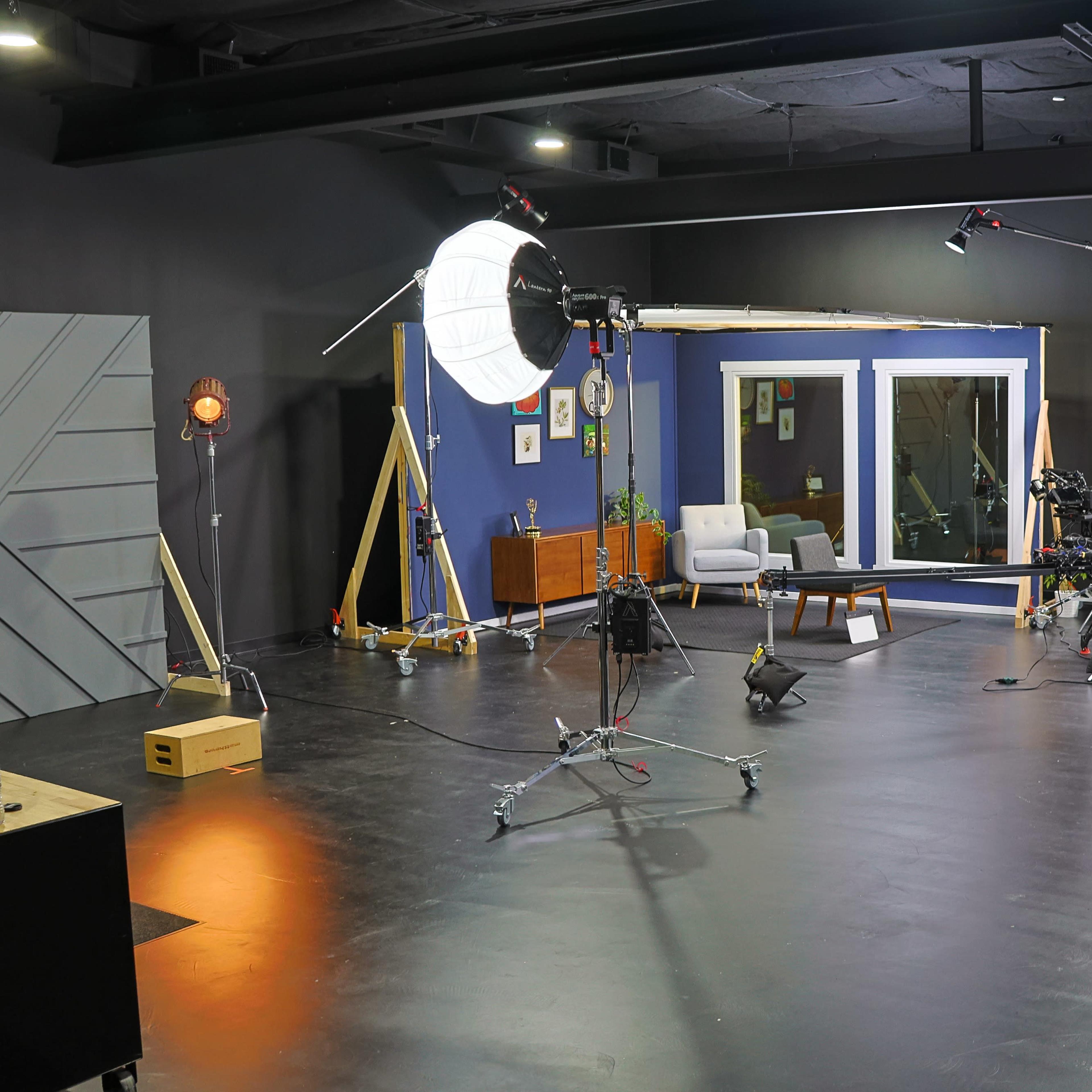 A photography studio is set up with various lighting equipment, cameras, and a staged area featuring a chair and a window.