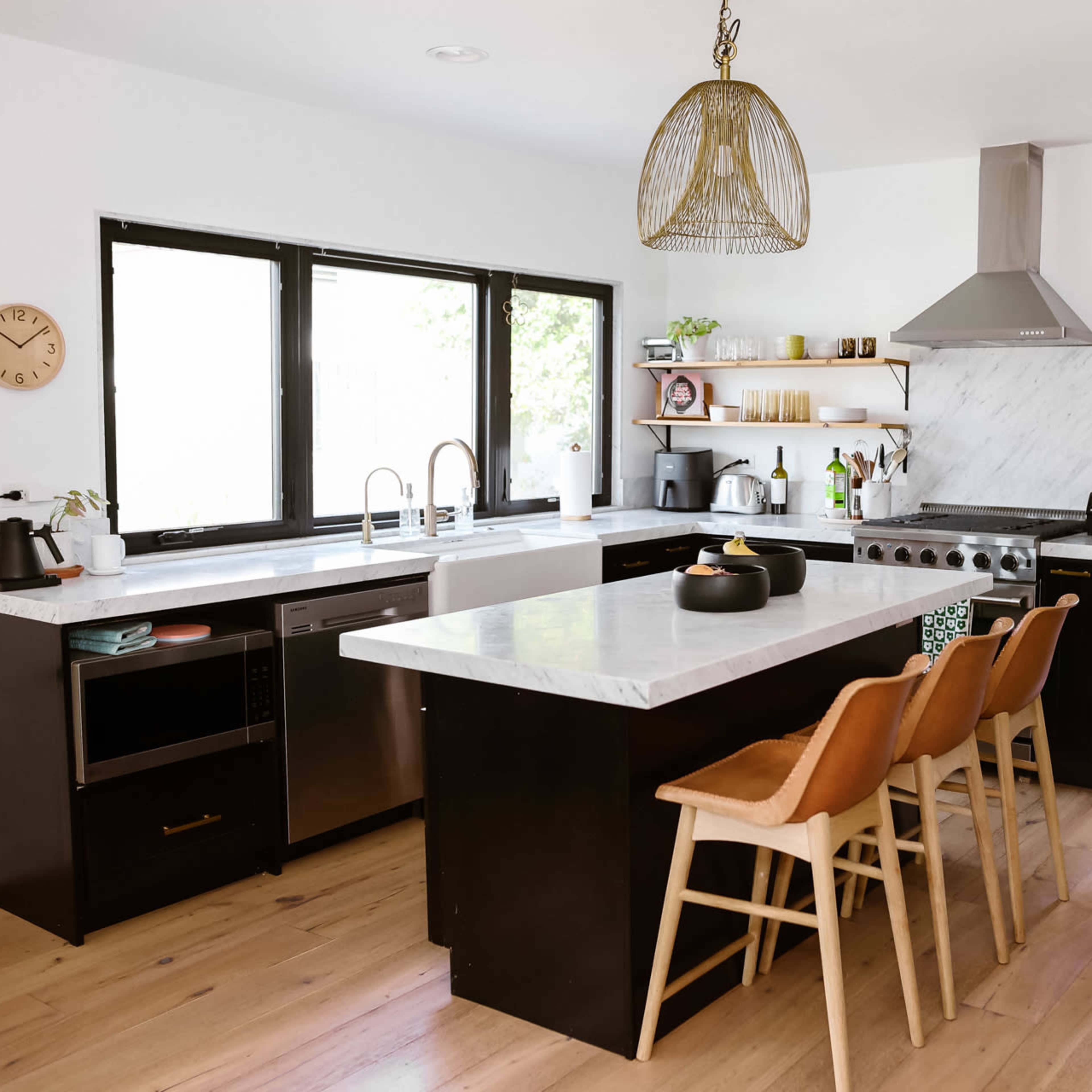A modern kitchen features a white marble countertop, black cabinetry, and wooden bar stools, with large windows allowing natural light to fill the space.