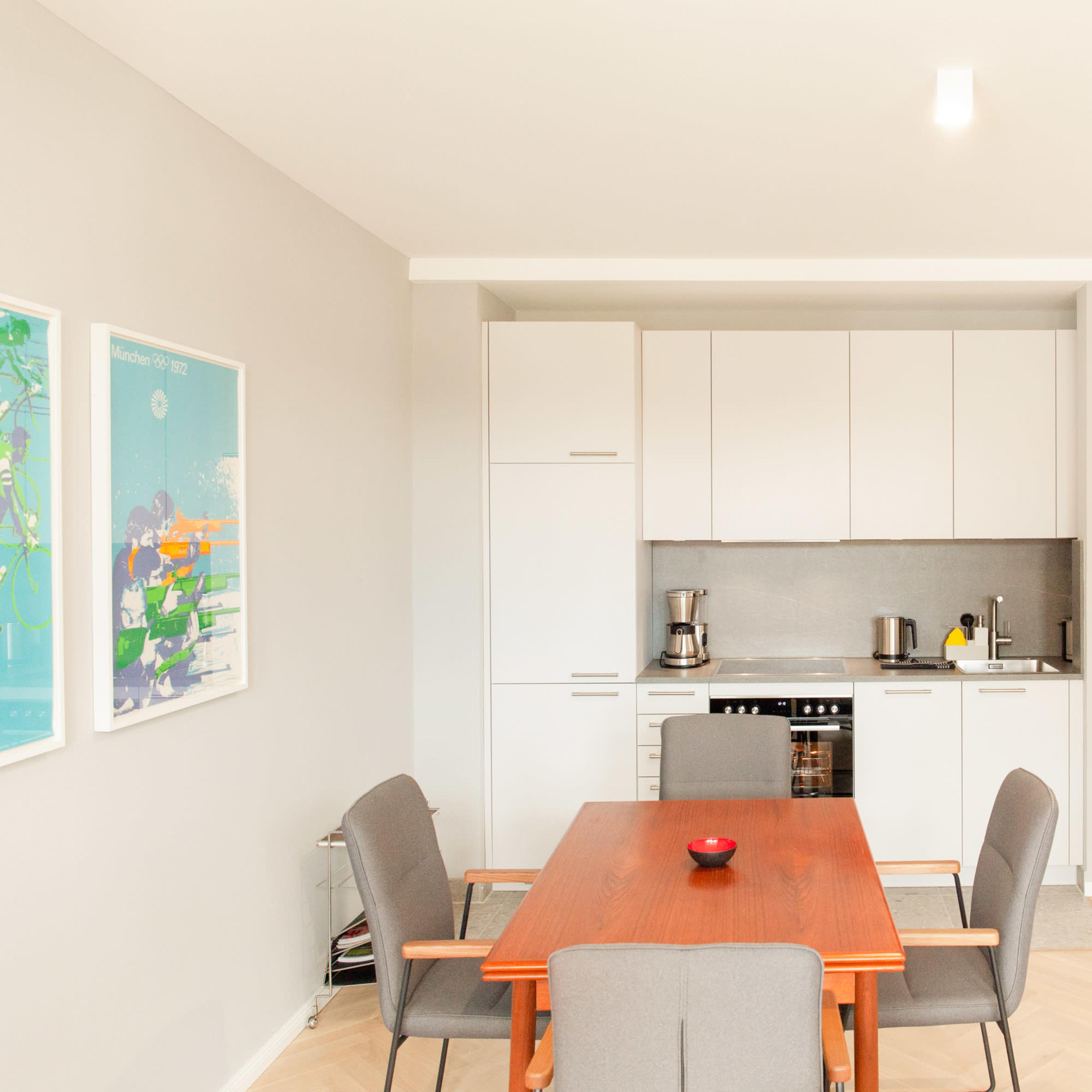 A modern kitchen and dining area feature a wooden table with gray chairs, sleek cabinetry, and a door leading to another room.