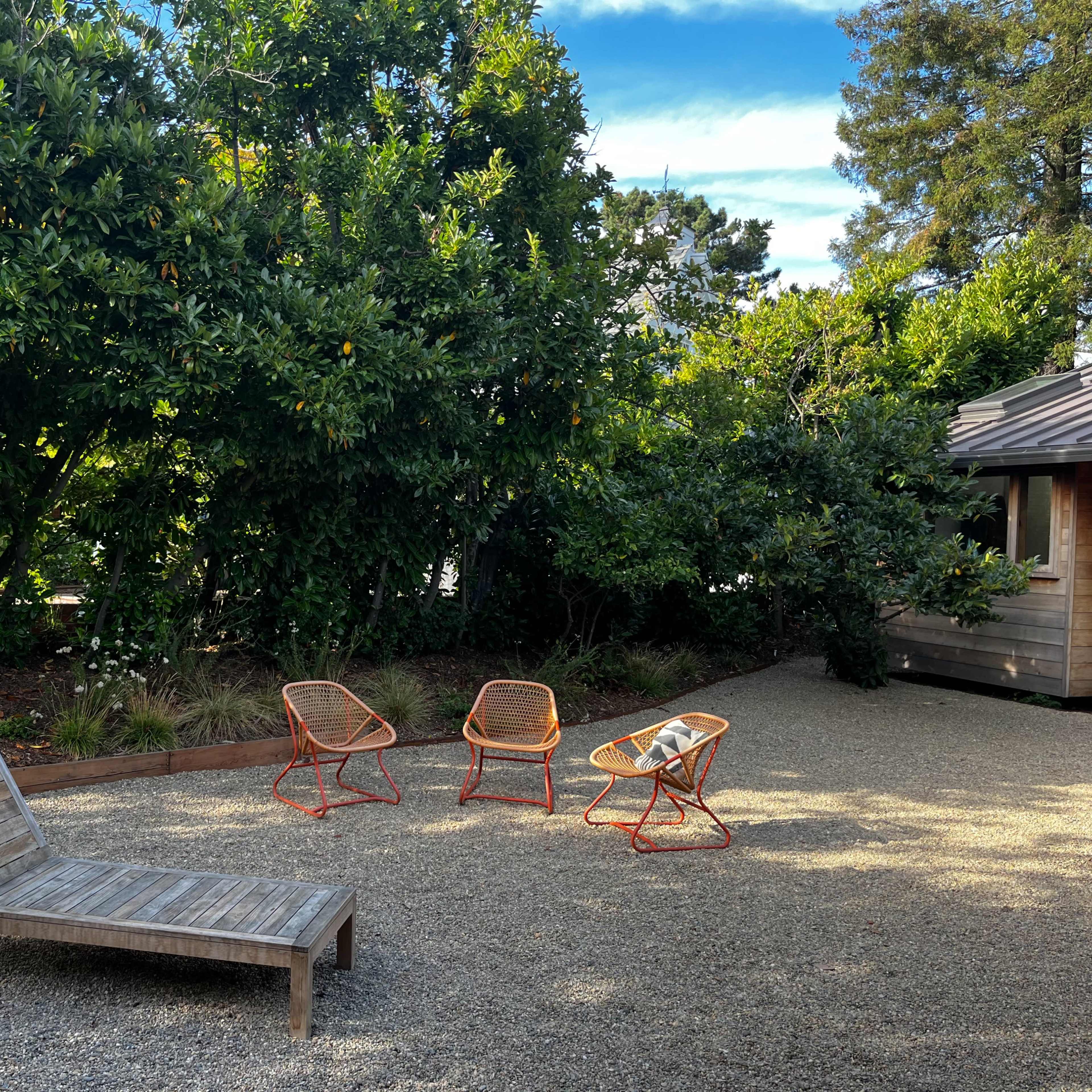 The scene features a wooden lounge chair and three colorful chairs arranged on a gravel surface surrounded by lush greenery and a nearby wooden building.