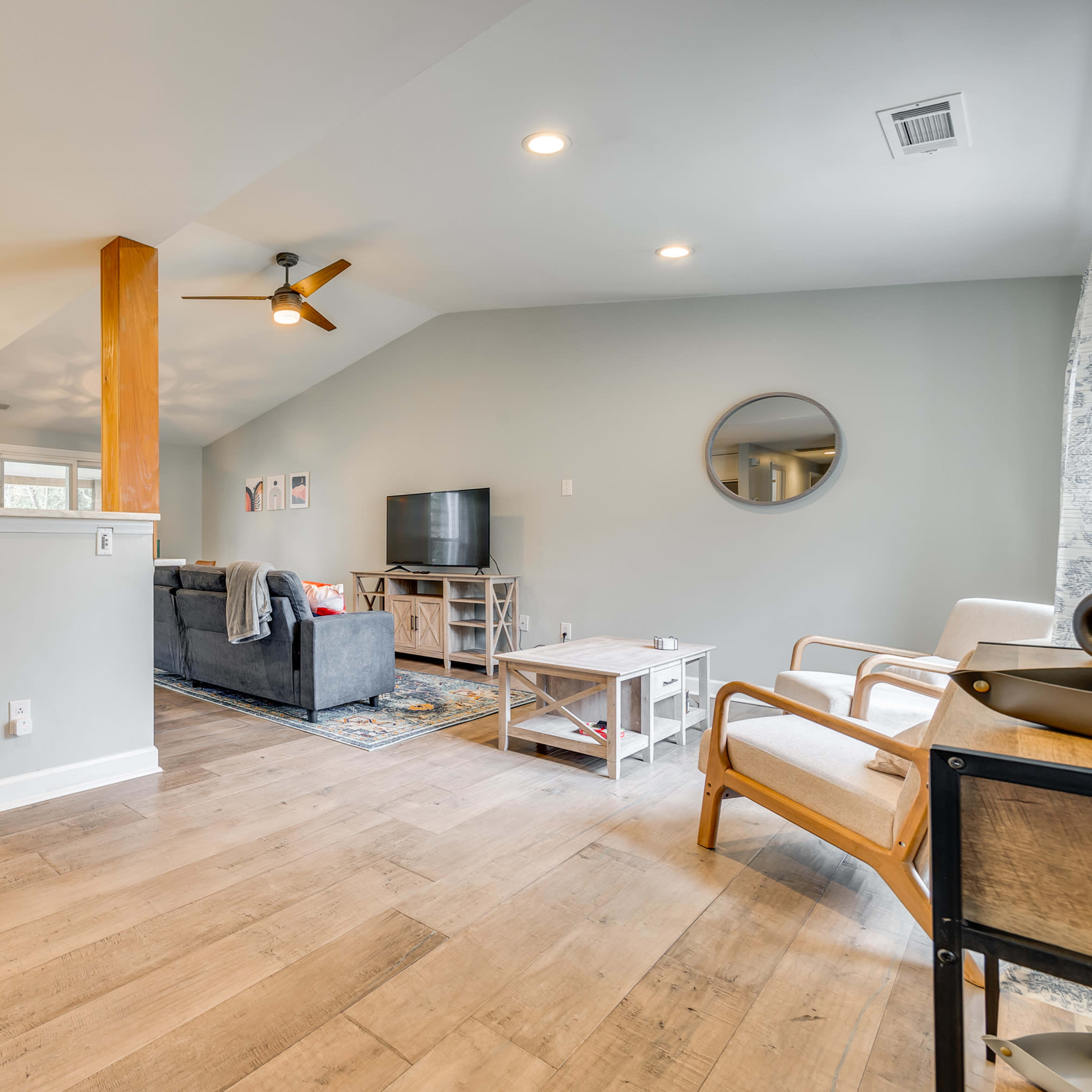 A spacious living area with a gray sofa, wooden flooring, and a ceiling fan, adjacent to a kitchen area with a large window and bright lighting.
