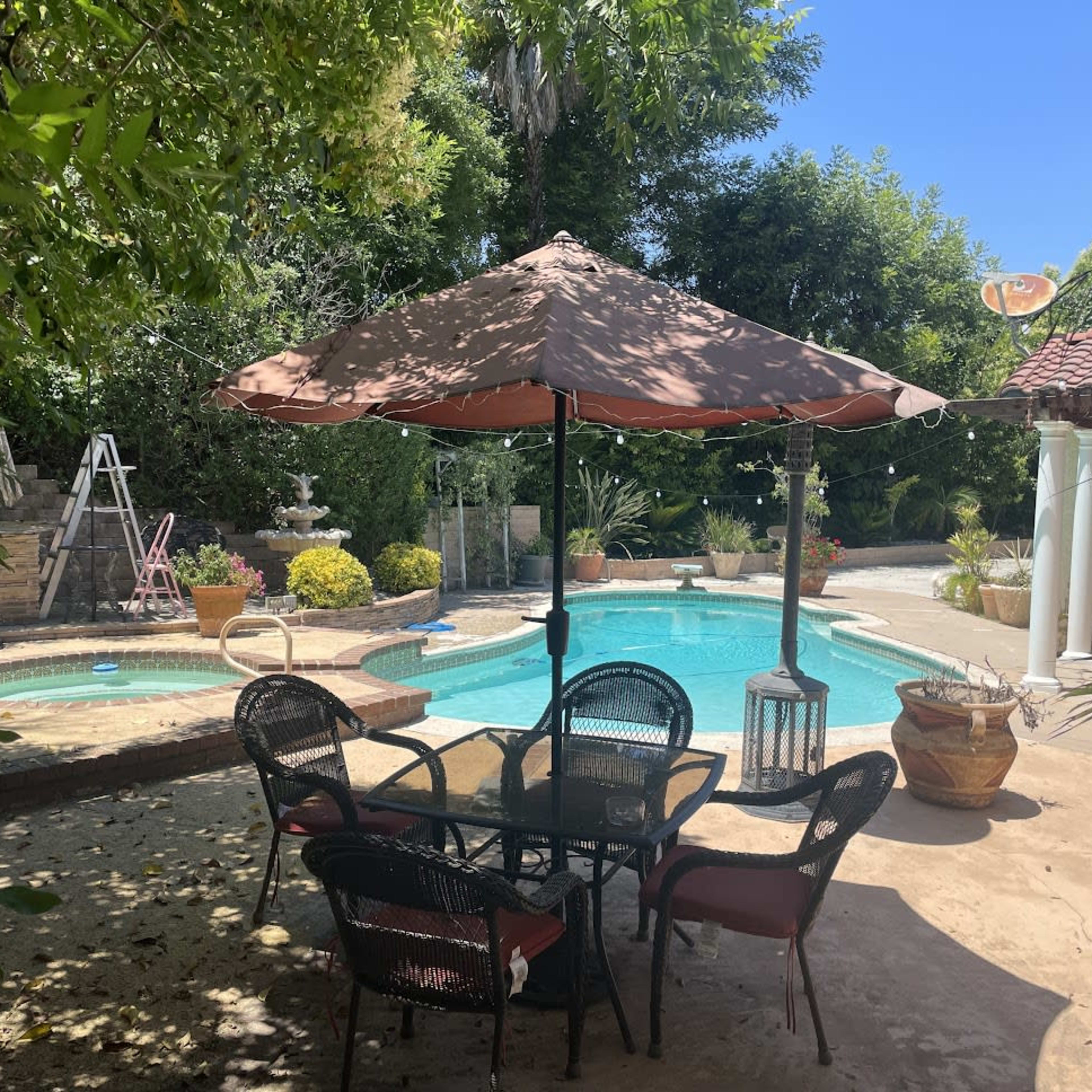A patio area with a table and chairs under an umbrella is positioned next to a swimming pool surrounded by greenery and decorative plants.
