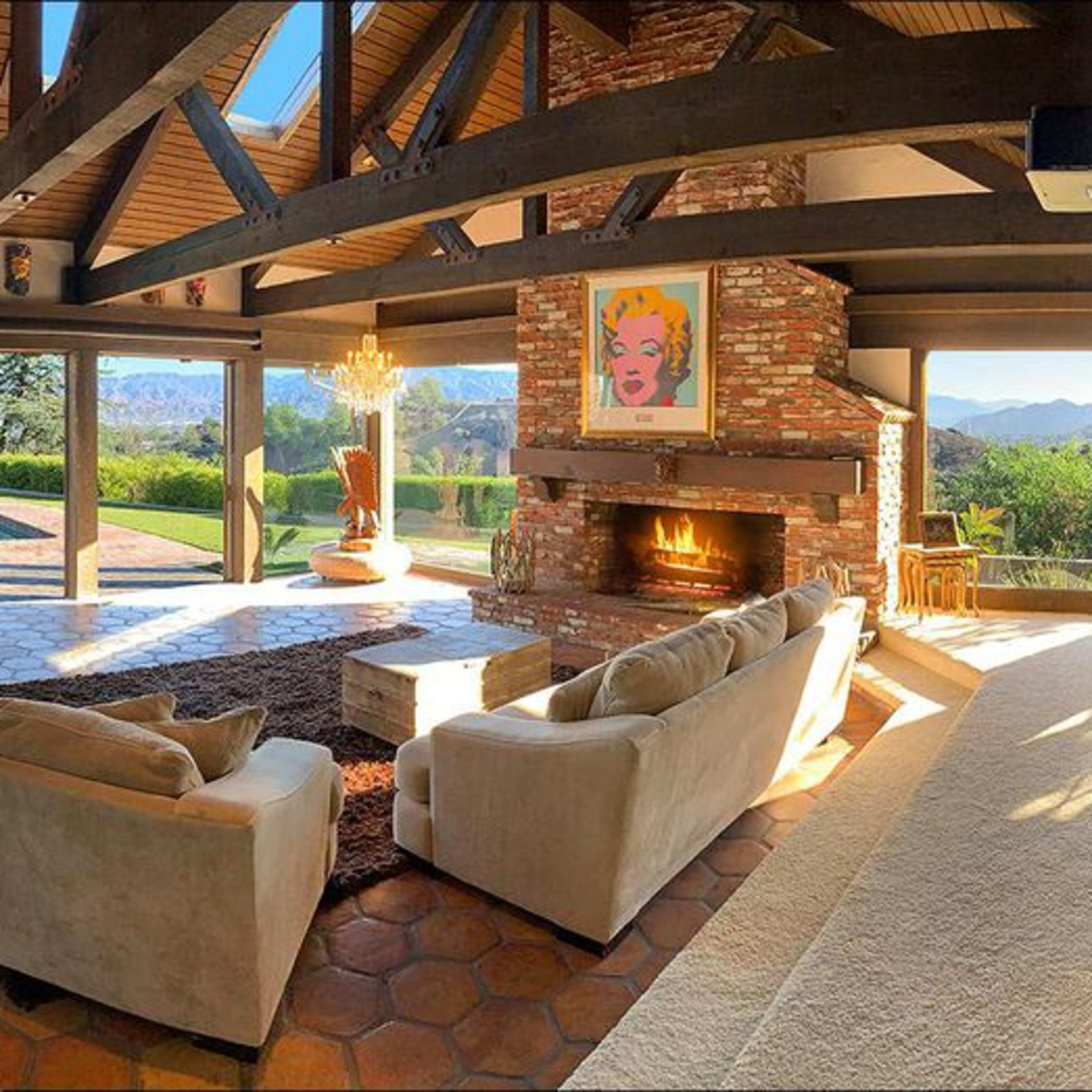 A spacious living room with a stone fireplace, large windows overlooking a landscape, and modern furniture arranged around a central coffee table.
