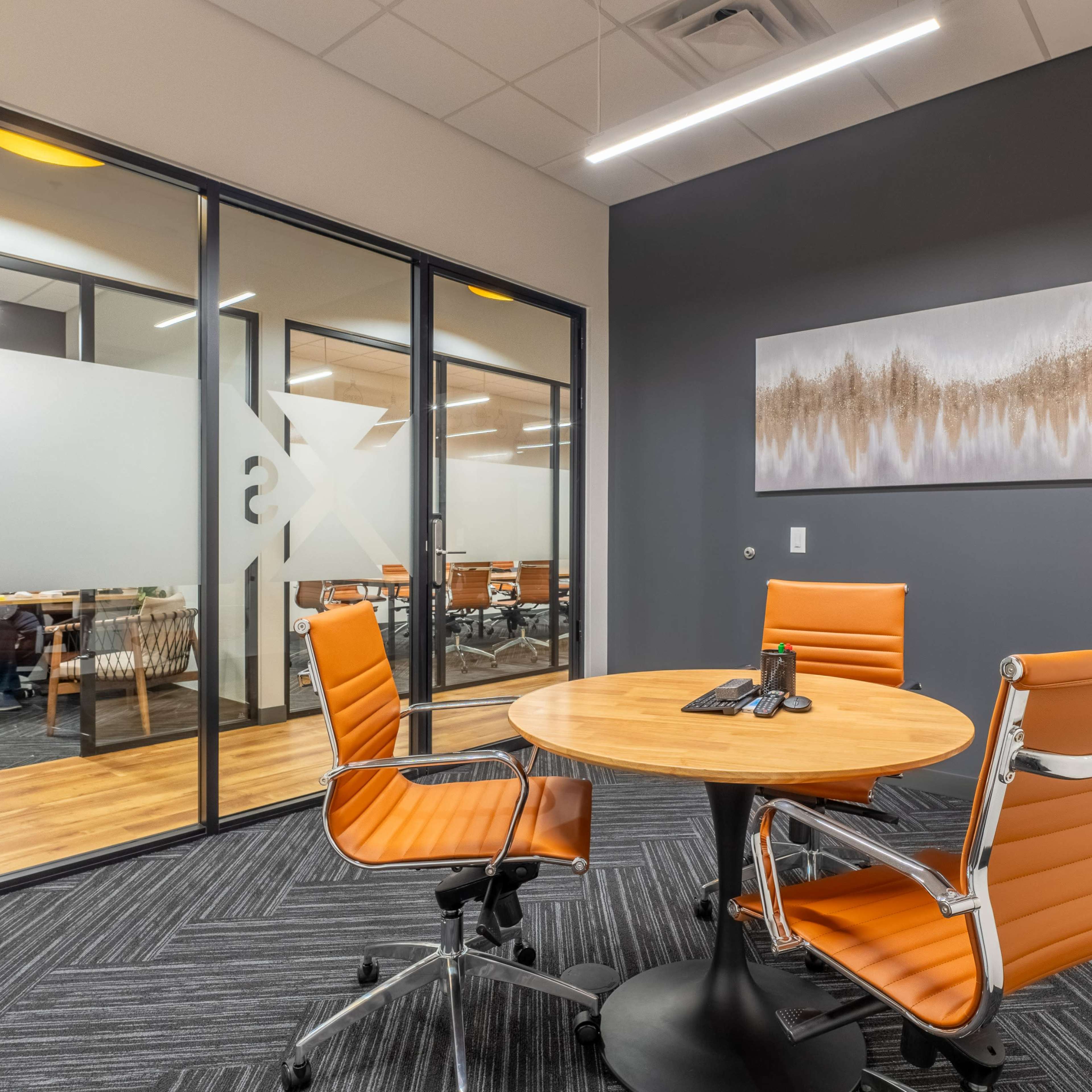 A modern office meeting space features a round wooden table surrounded by three orange chairs, with a large glass partition displaying additional workspaces in the background.