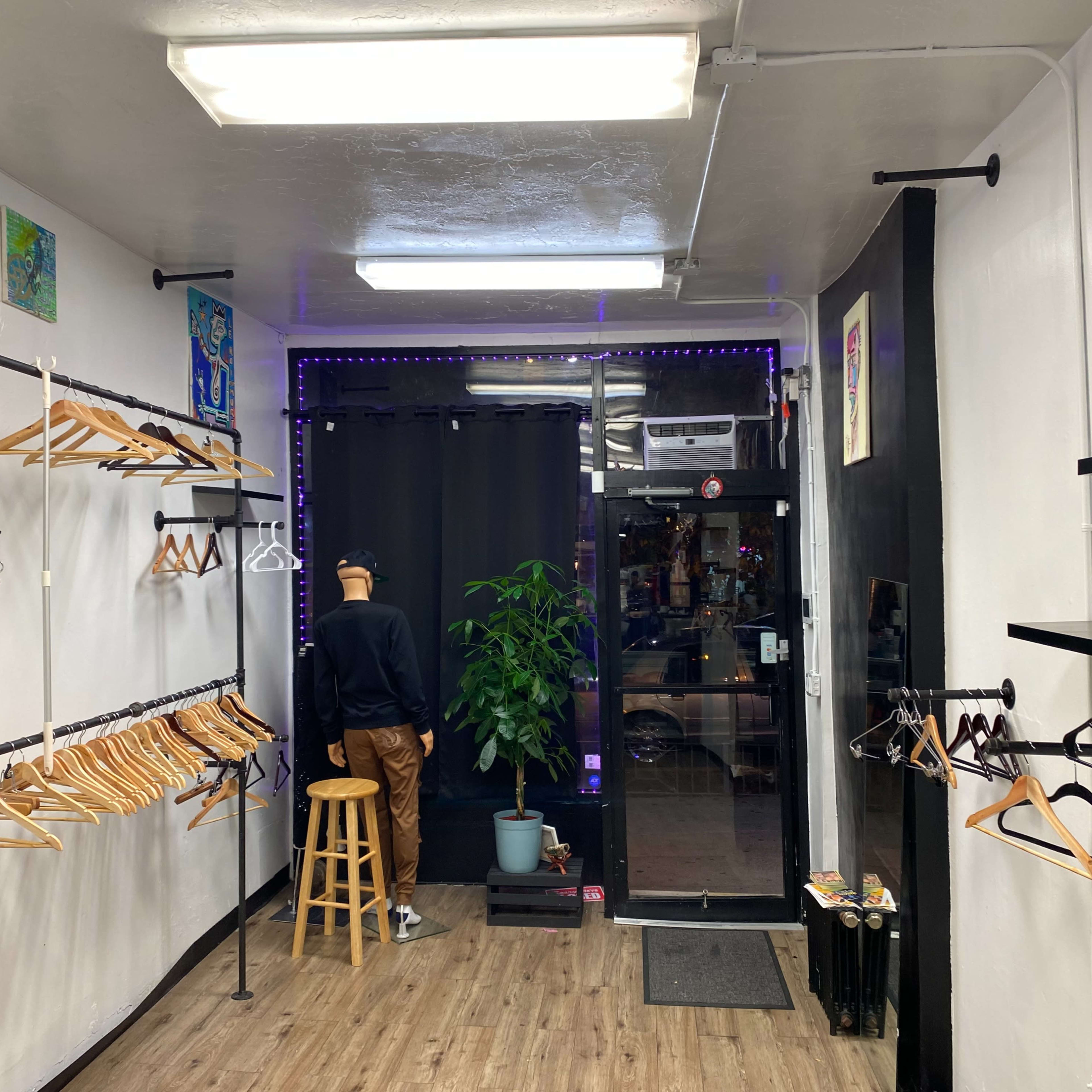 A clothing store interior with wooden hangers on metal racks, a stool, a plant in a pot, and a mannequin facing a curtained entrance.