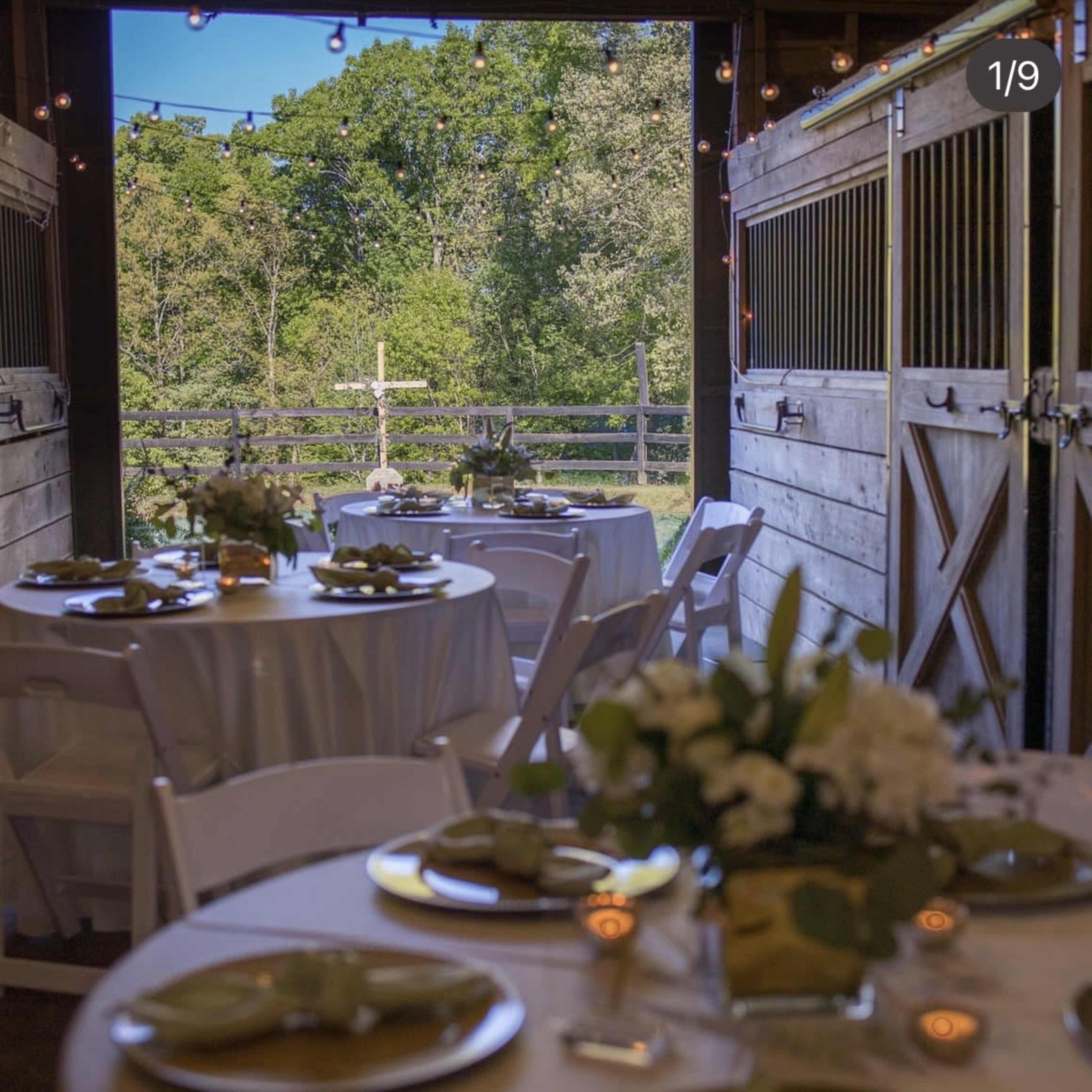 A barn interior features elegantly set tables with white tablecloths and floral centerpieces, opening to a view of greenery through the stable doors.