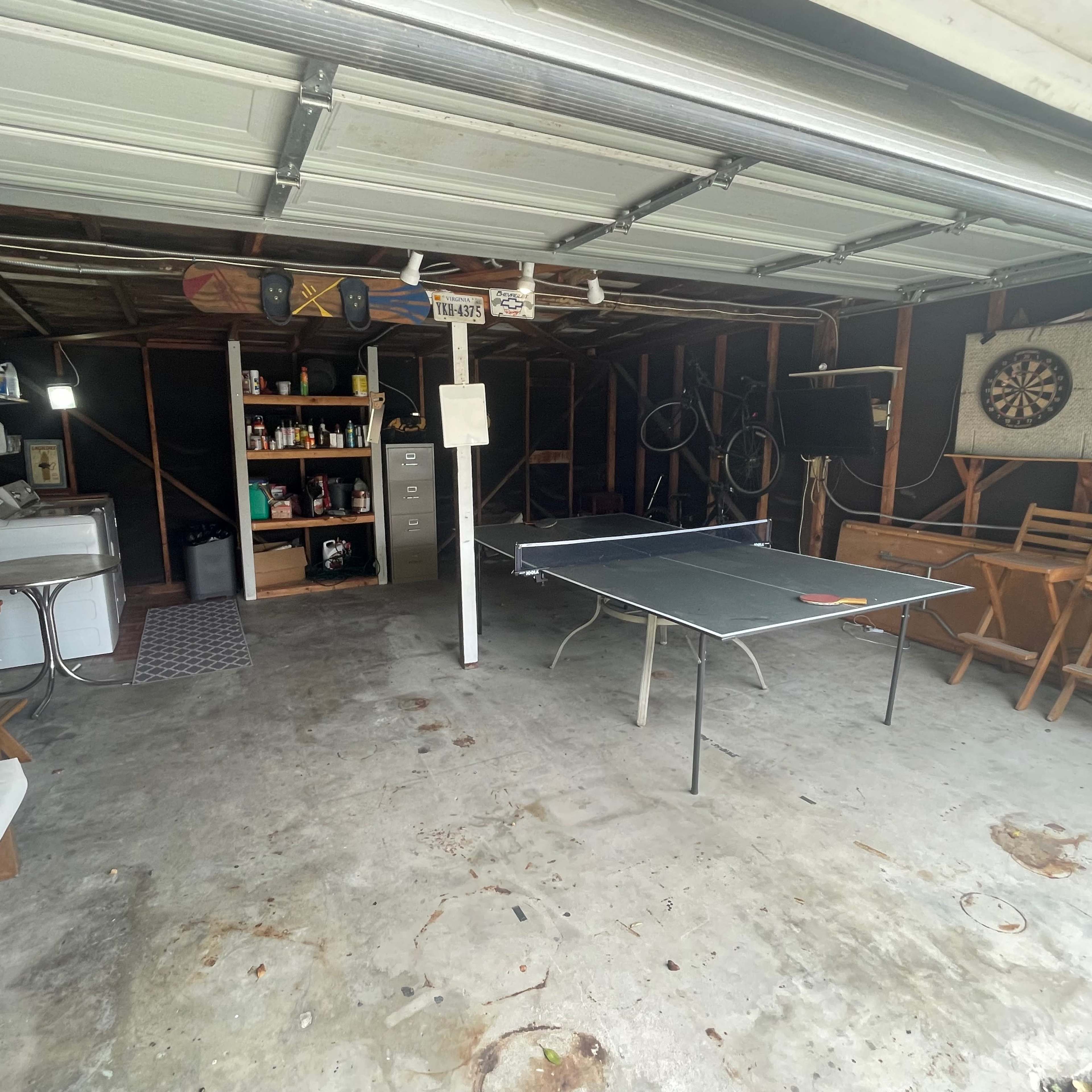 A spacious garage with a ping pong table, shelves of supplies, and various furniture including chairs and a washing machine.