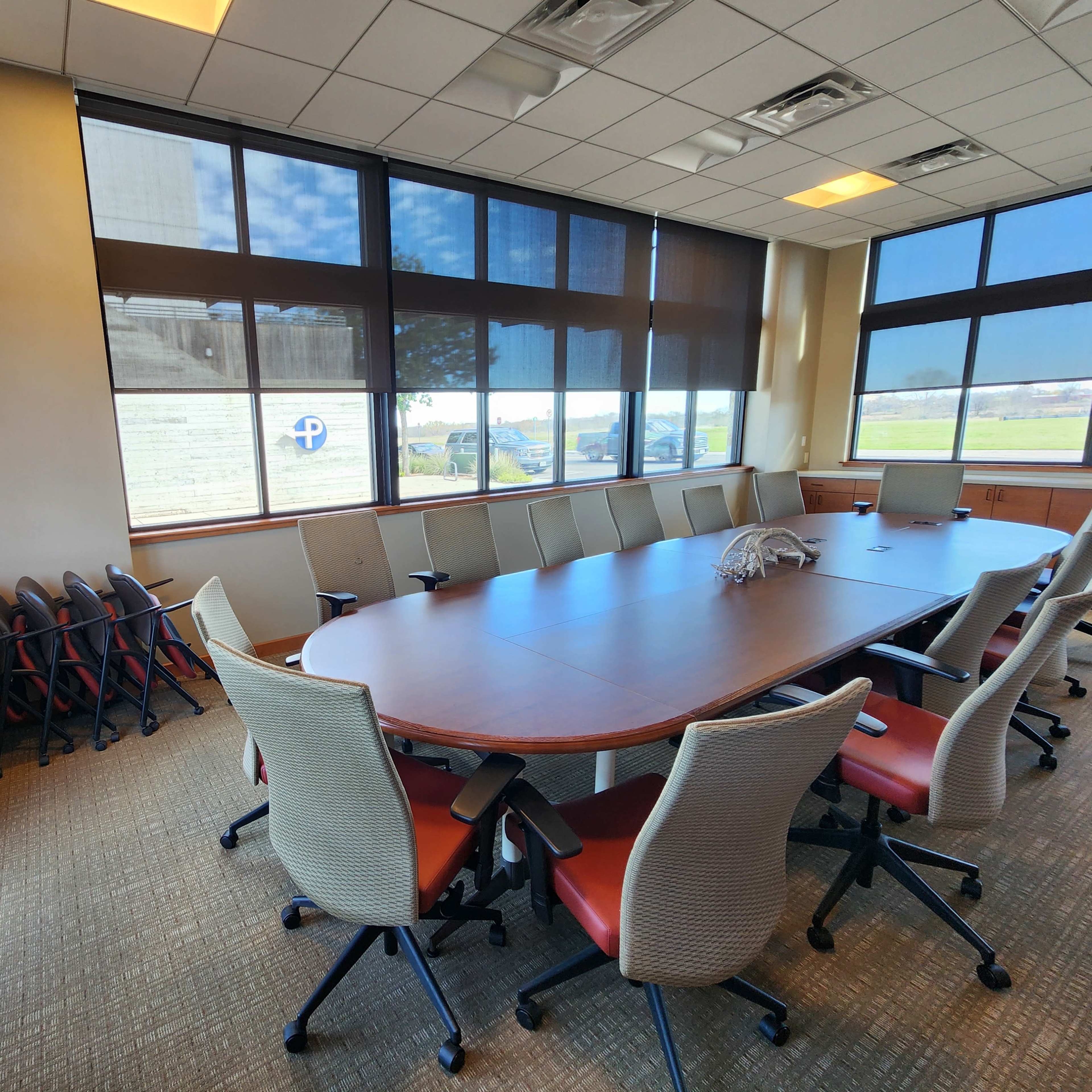 A conference room with a large oval table, rolling chairs, and windows providing natural light.