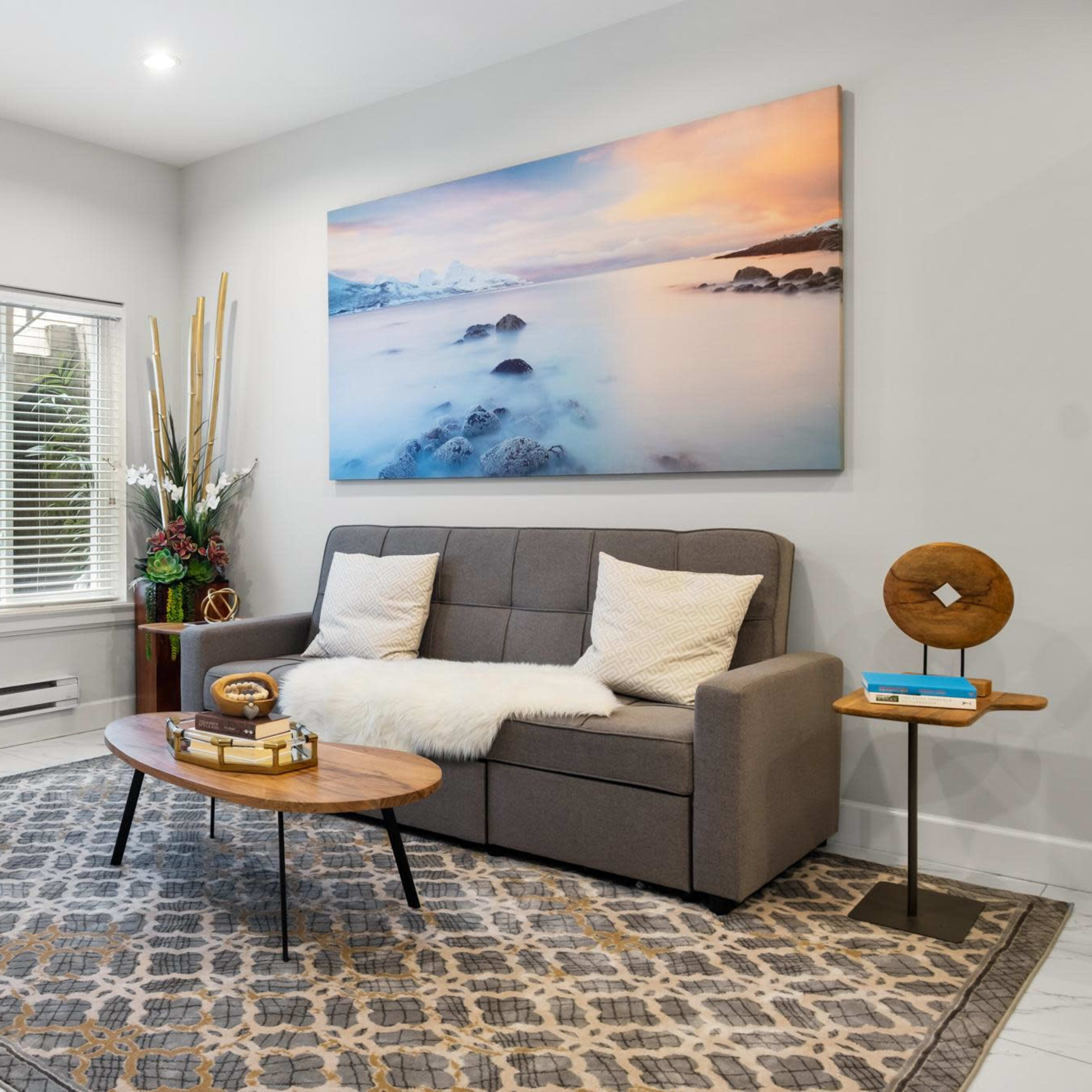 A modern living room featuring a gray sofa, a round wooden coffee table, and a light-colored armchair, with a large landscape painting on the wall and a staircase visible in the background.