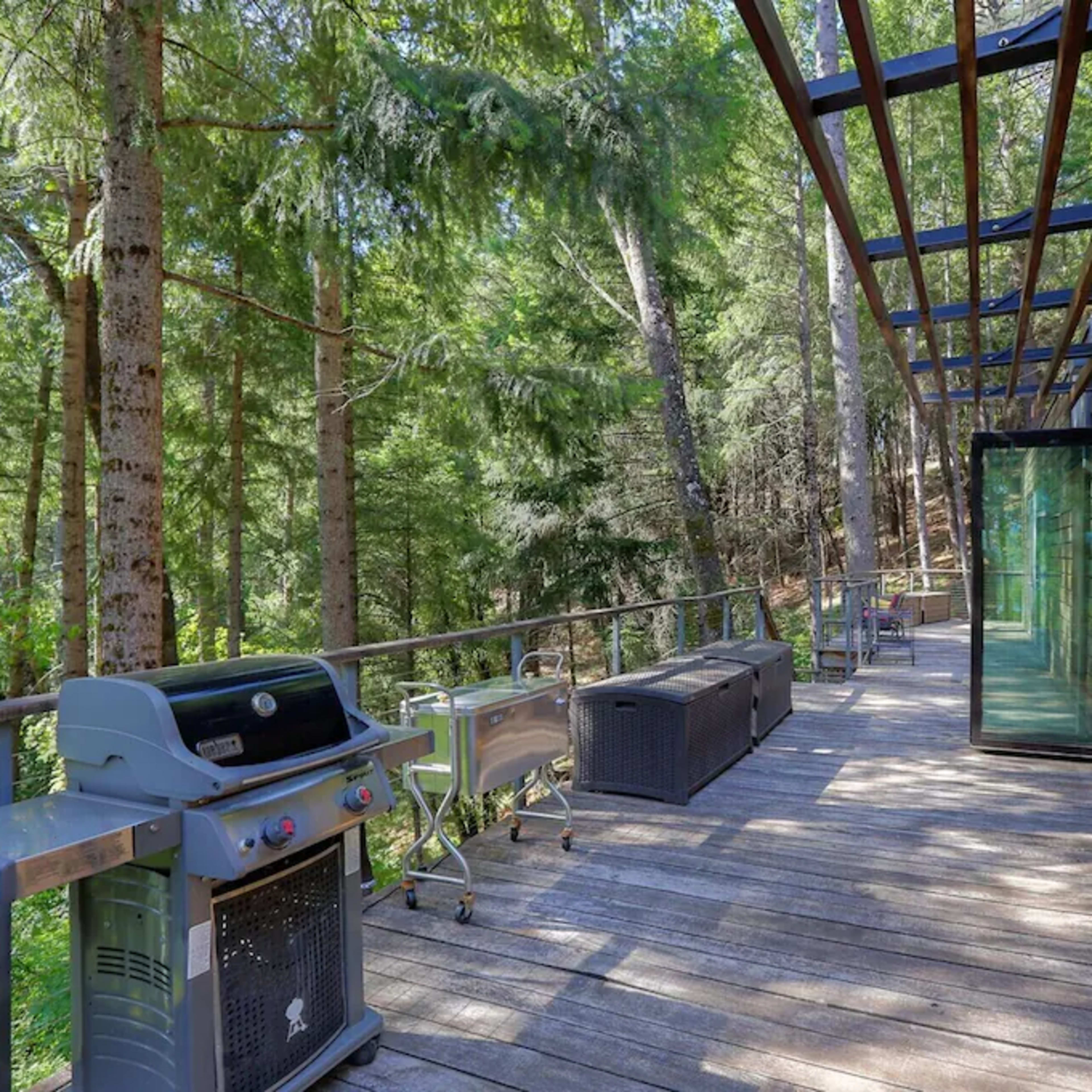 A wooden deck with a barbecue grill overlooks a wooded area, featuring lounge furniture and large glass doors leading into a modern interior.