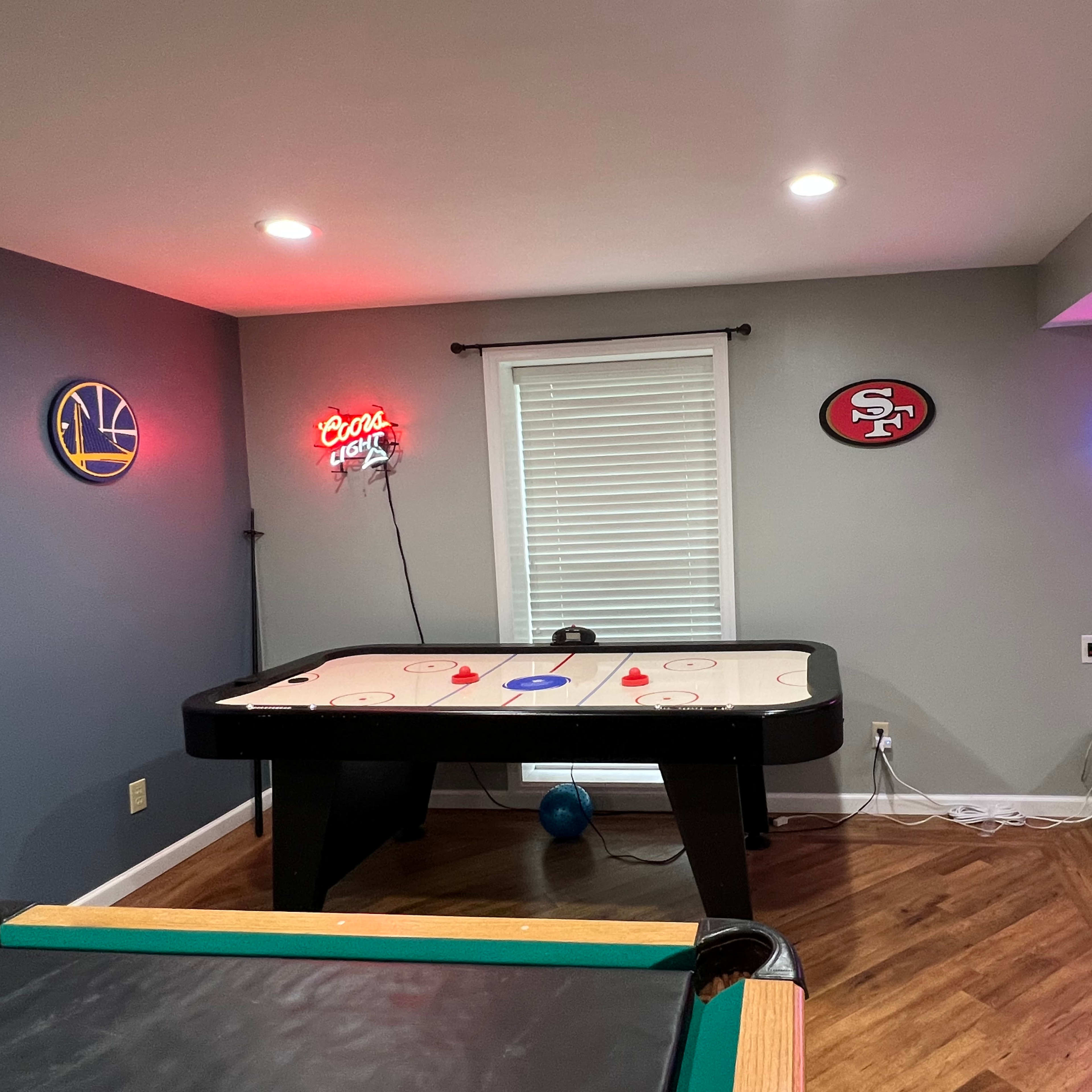 A game room featuring an air hockey table, a pool table, and neon signs indicating "SUBS" and a logo, with wood flooring and a window covered by blinds.