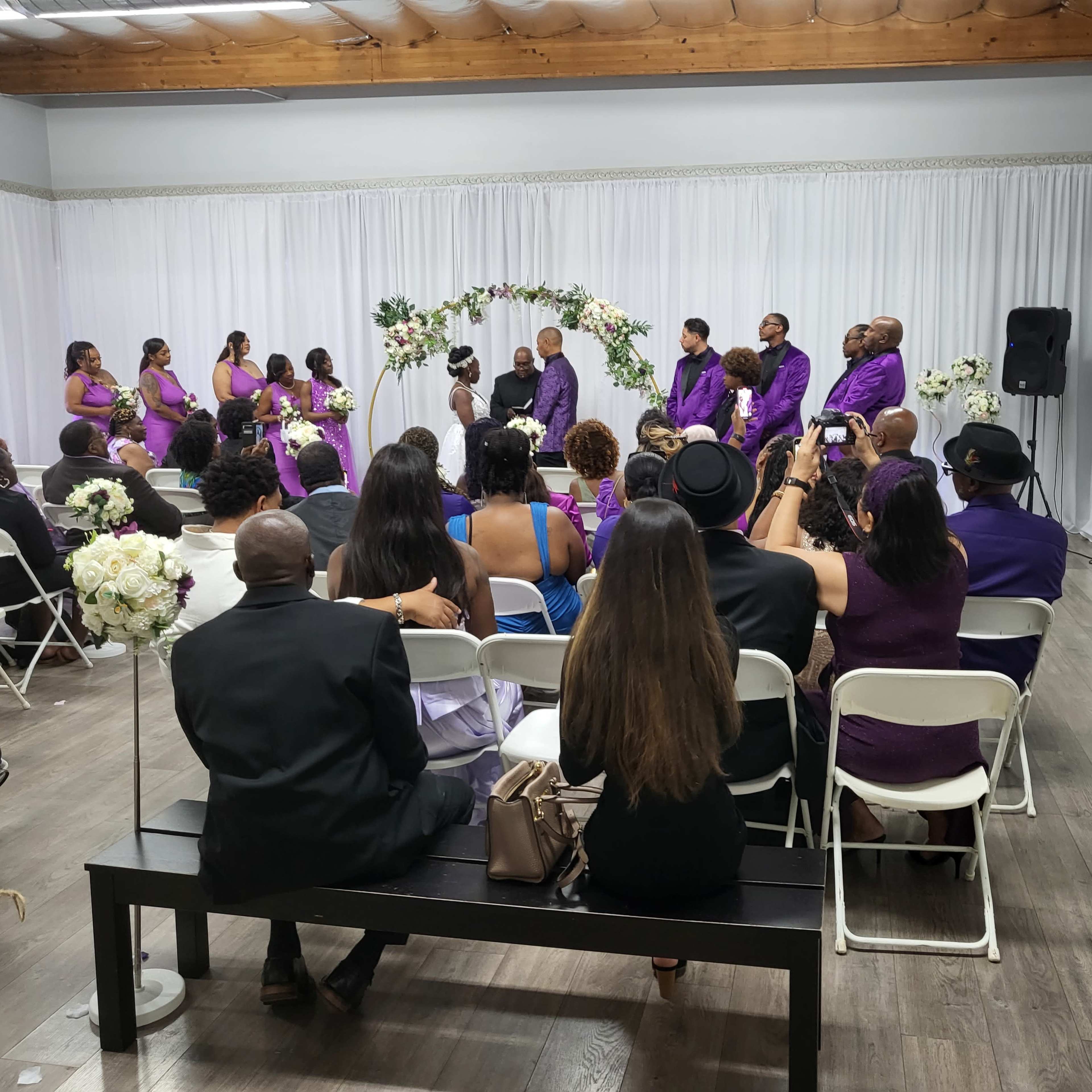 A wedding ceremony is taking place indoors, featuring a couple standing at an altar surrounded by attendants and seated guests.