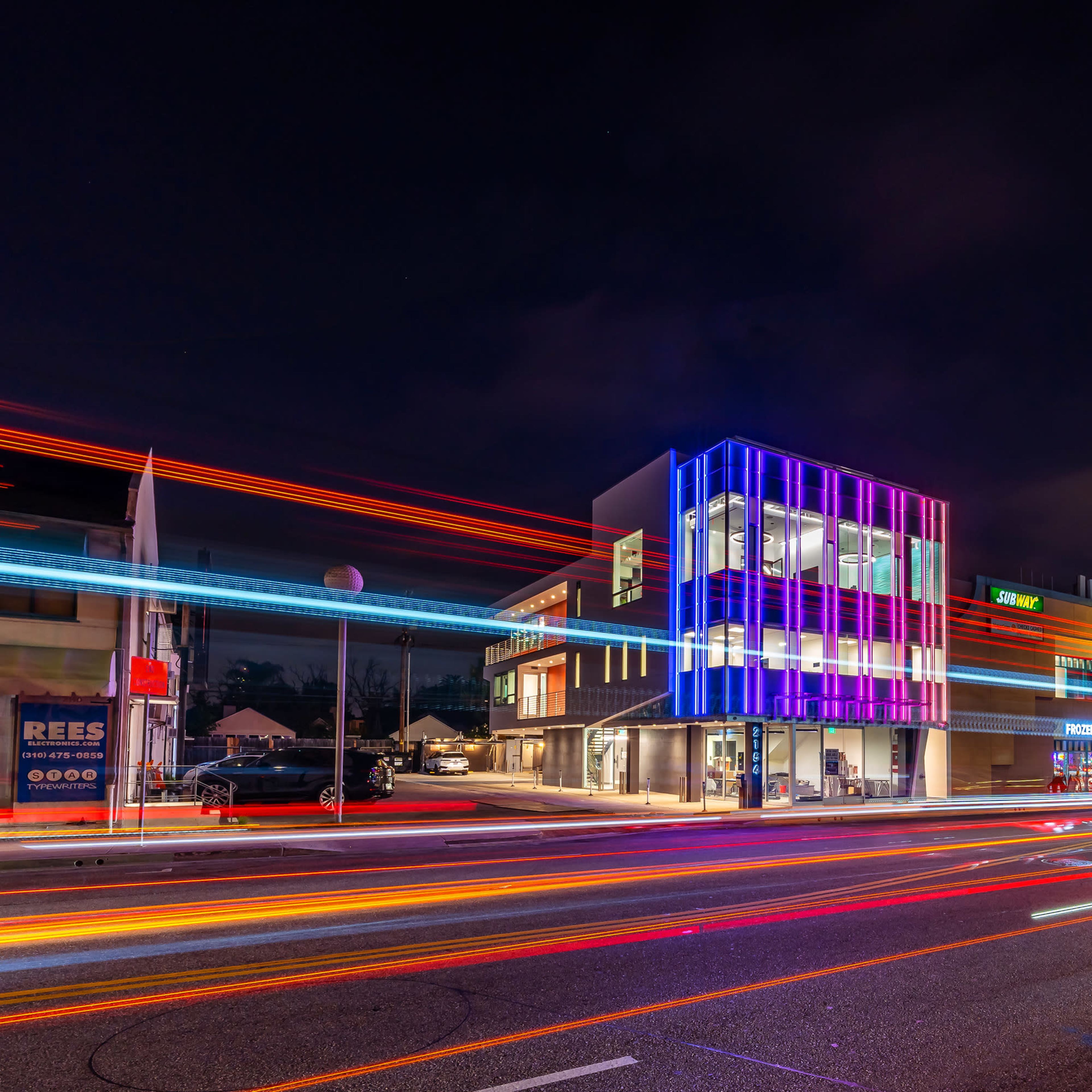 A modern building with colorful light accents stands beside a busy road at night, with light trails from passing vehicles creating a dynamic effect.