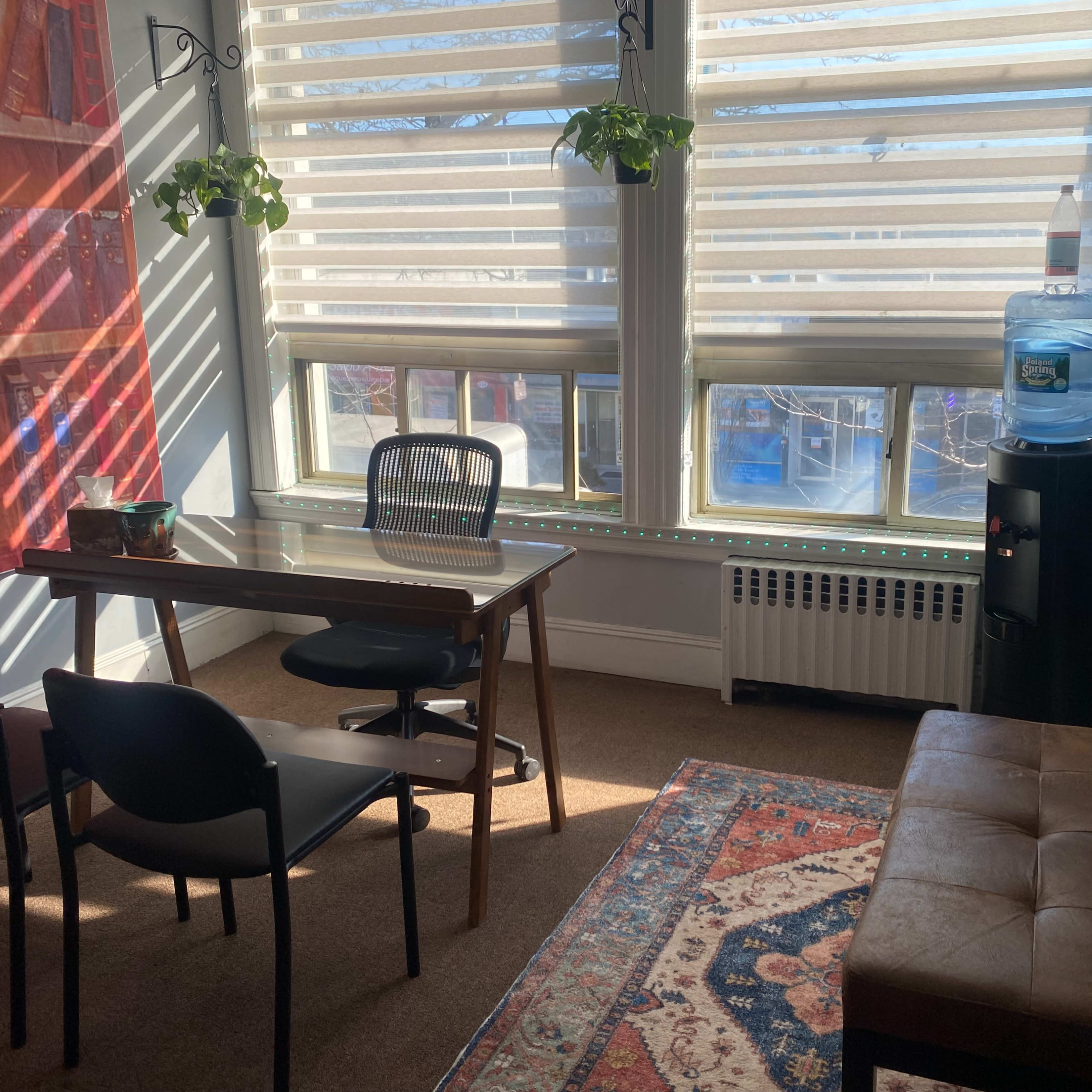 A small office space features a wooden table, black chairs, a water cooler, and large windows with blinds, illuminated by natural light.