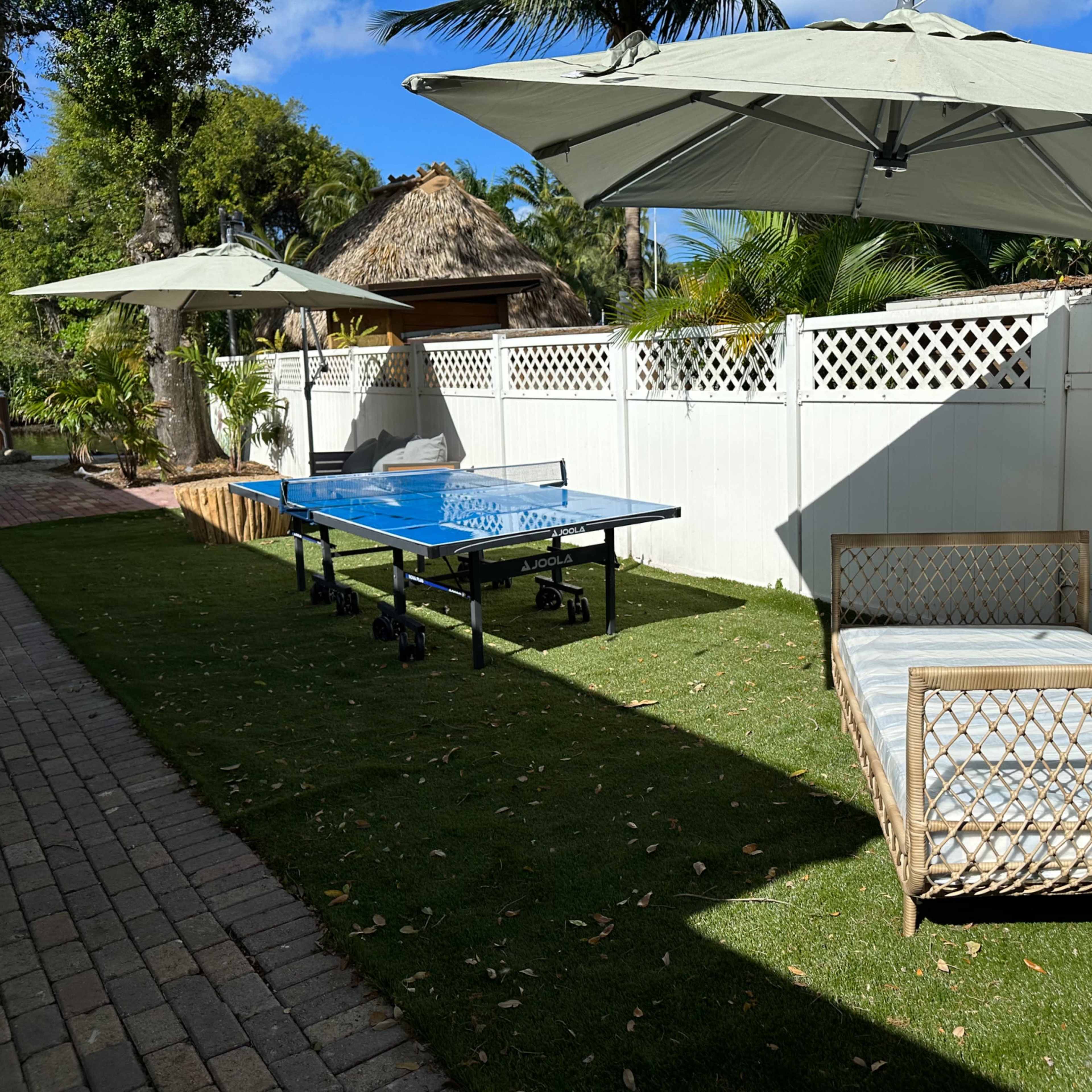 A backyard area with a ping pong table, a sunbed, and large umbrellas on a grassy lawn beside a white fence.