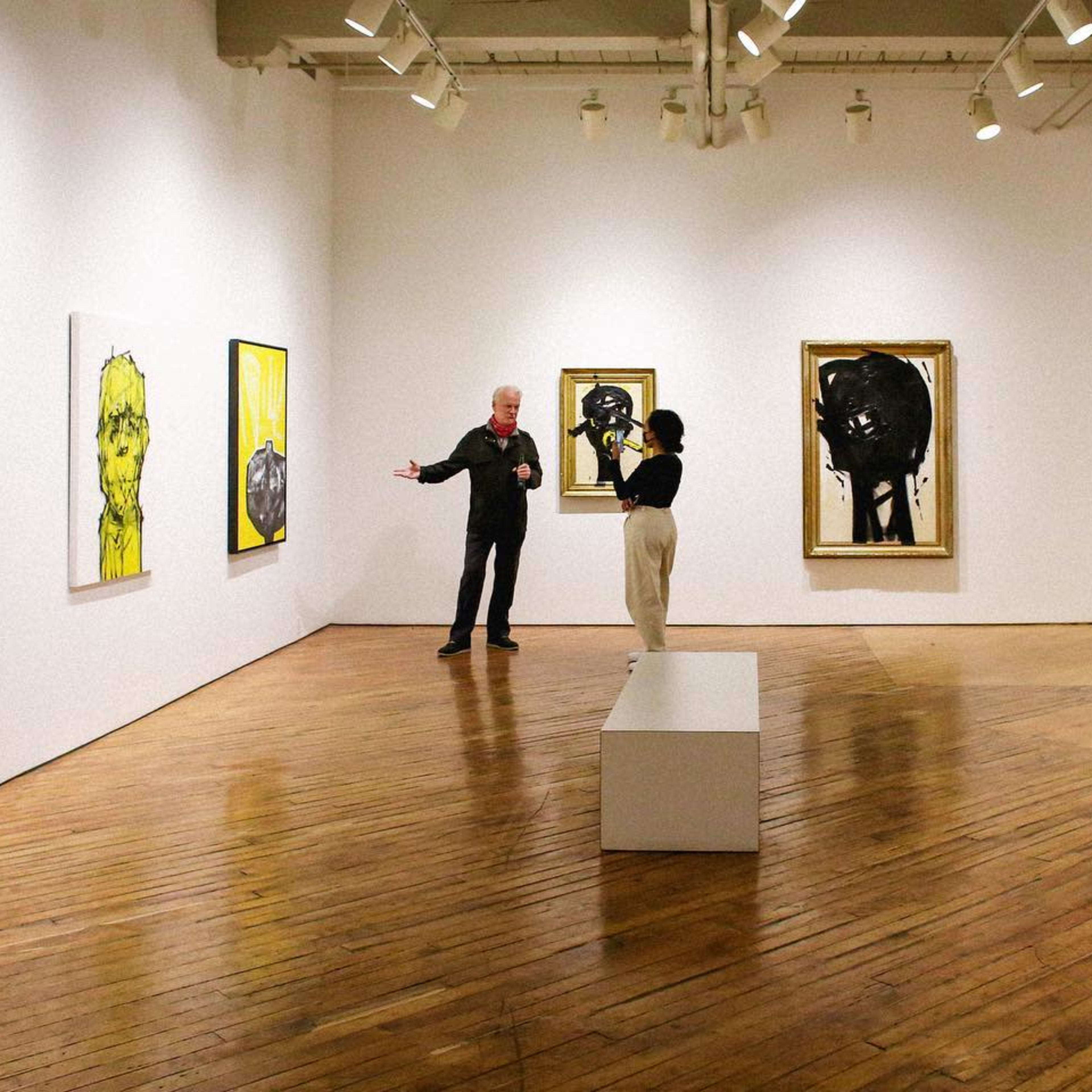 A man and a woman are discussing art in a gallery filled with large paintings on white walls and a wooden floor.