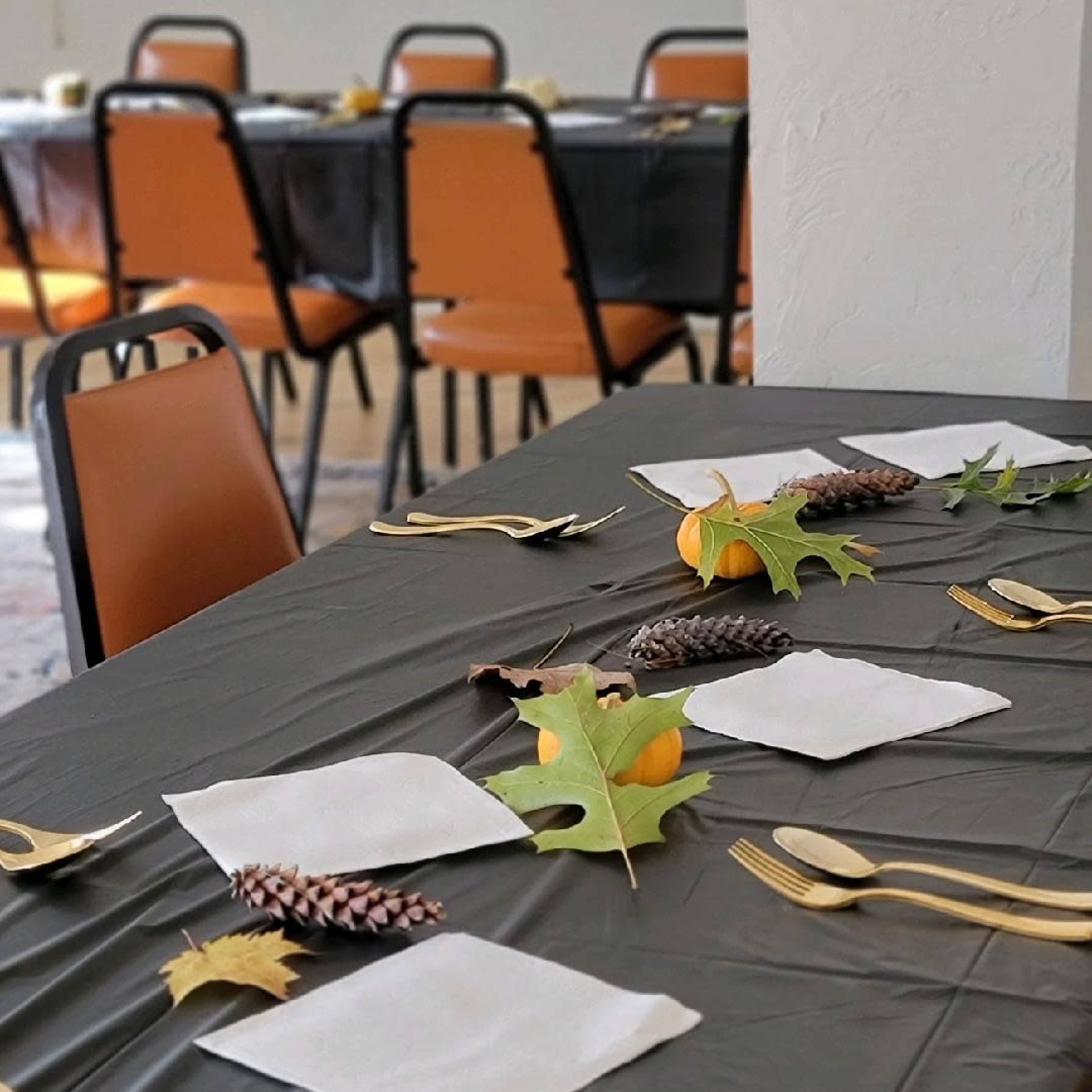 A dining table is set with black tablecloths, white napkins, pinecones, and autumn leaves, with chairs arranged around it in a bright room.