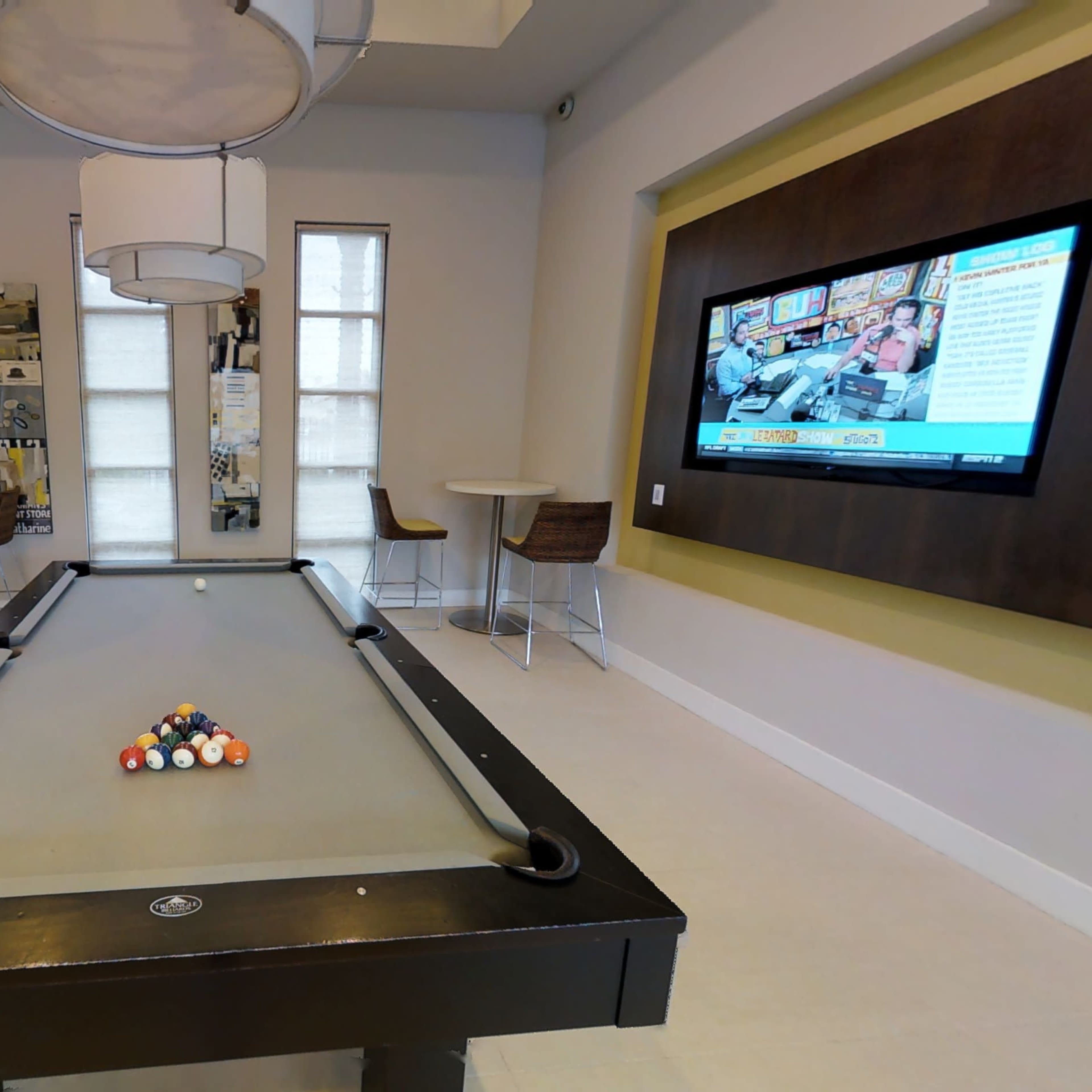 A game room featuring a pool table with a full rack of balls, two bar-style tables, and a television mounted on the wall.