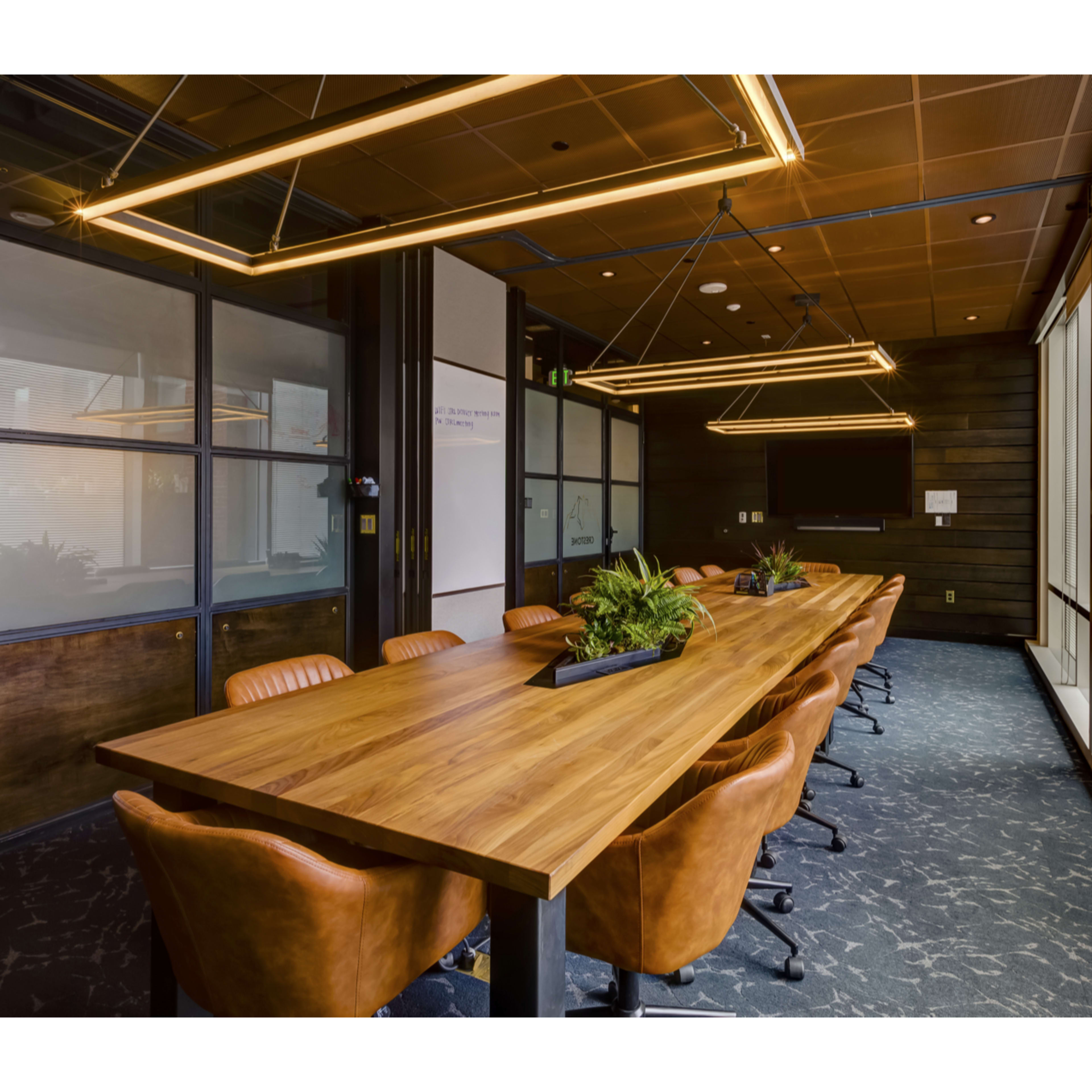 A long wooden conference table with leather chairs is placed in a modern office meeting room featuring large windows and suspended light fixtures.