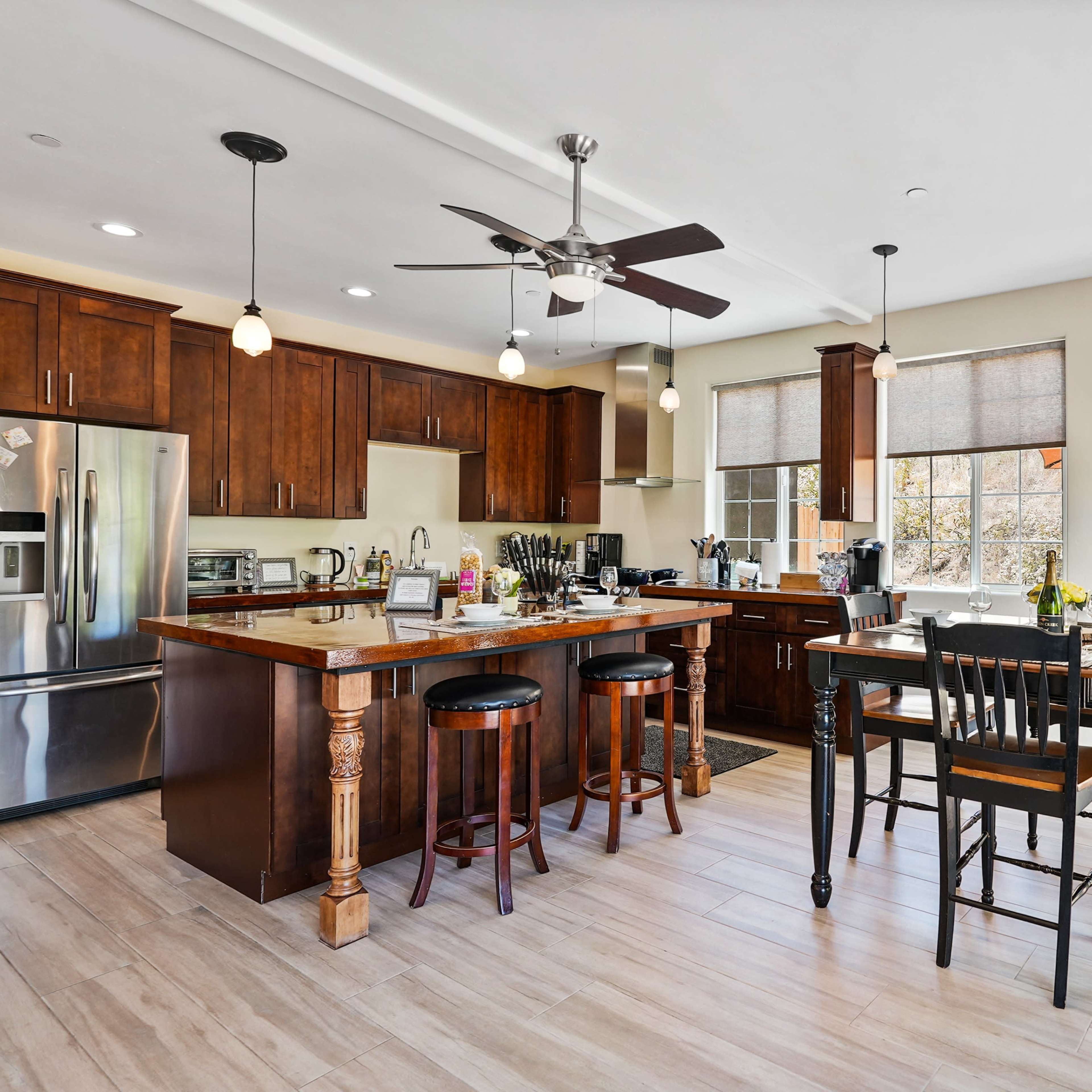 A modern kitchen features wooden cabinets, a central island with bar stools, and a dining table in front of large windows.