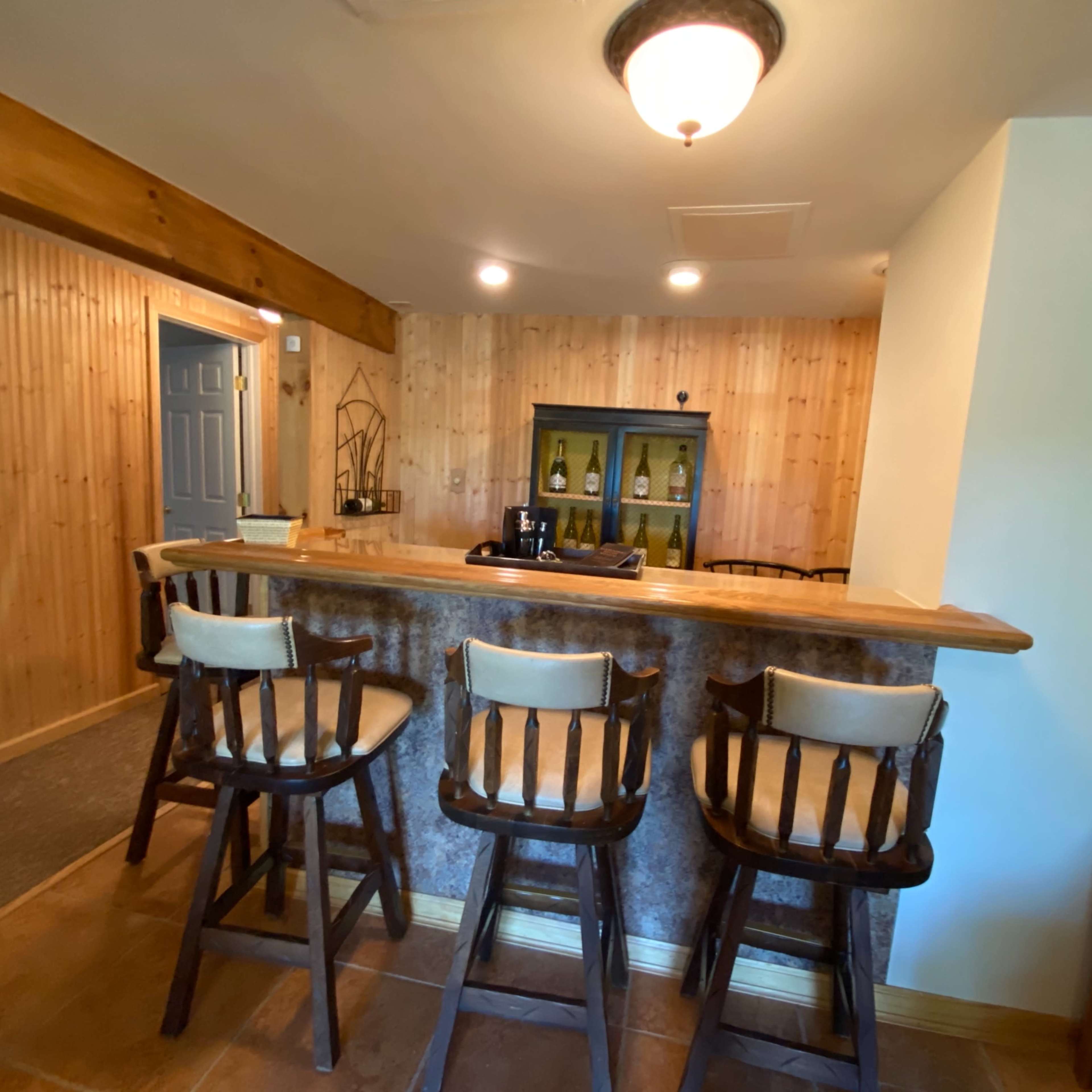 A wooden bar area with four high chairs and a wall-mounted cabinet displaying bottles, set against a backdrop of wooden paneling.