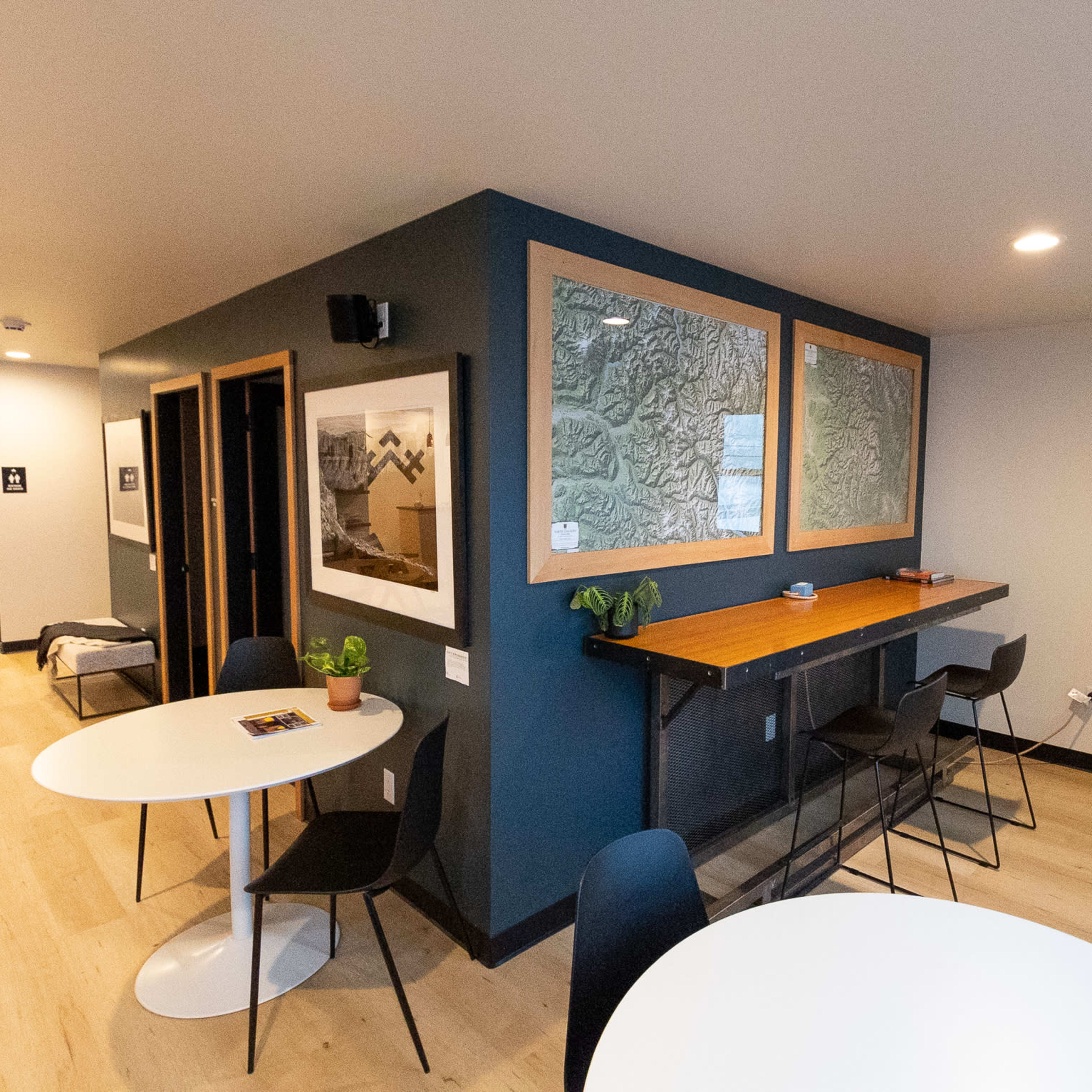 A modern interior space featuring a bar-height table with stools, two round white tables, and a combination of framed art and textured glass panels on the walls.