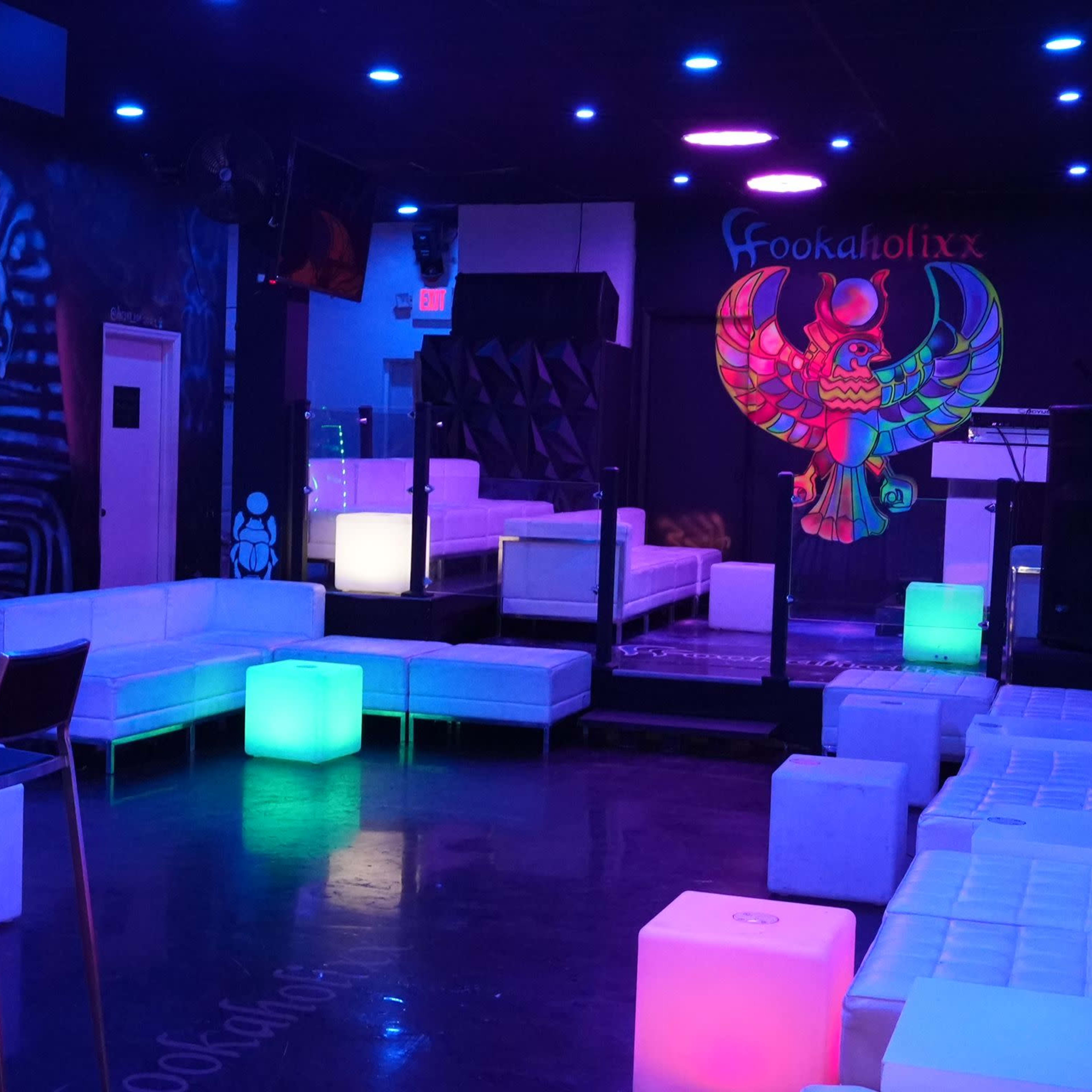 The interior of a vibrant nightclub features white seating, colorful illuminated tables, and large graffiti art on the walls.
