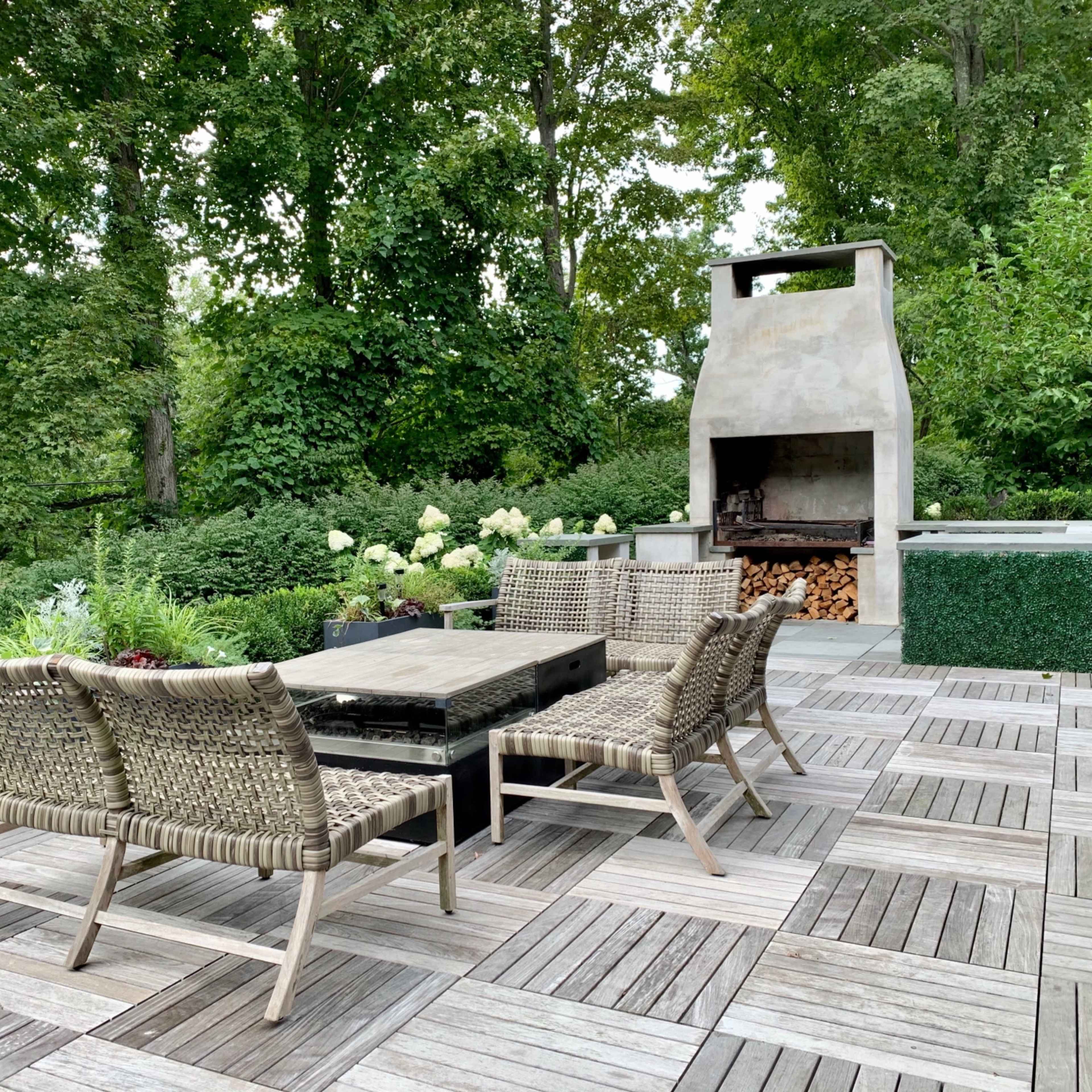 A landscaped outdoor patio area with a seating arrangement of woven chairs and a central table near a stone barbecue grill surrounded by greenery.