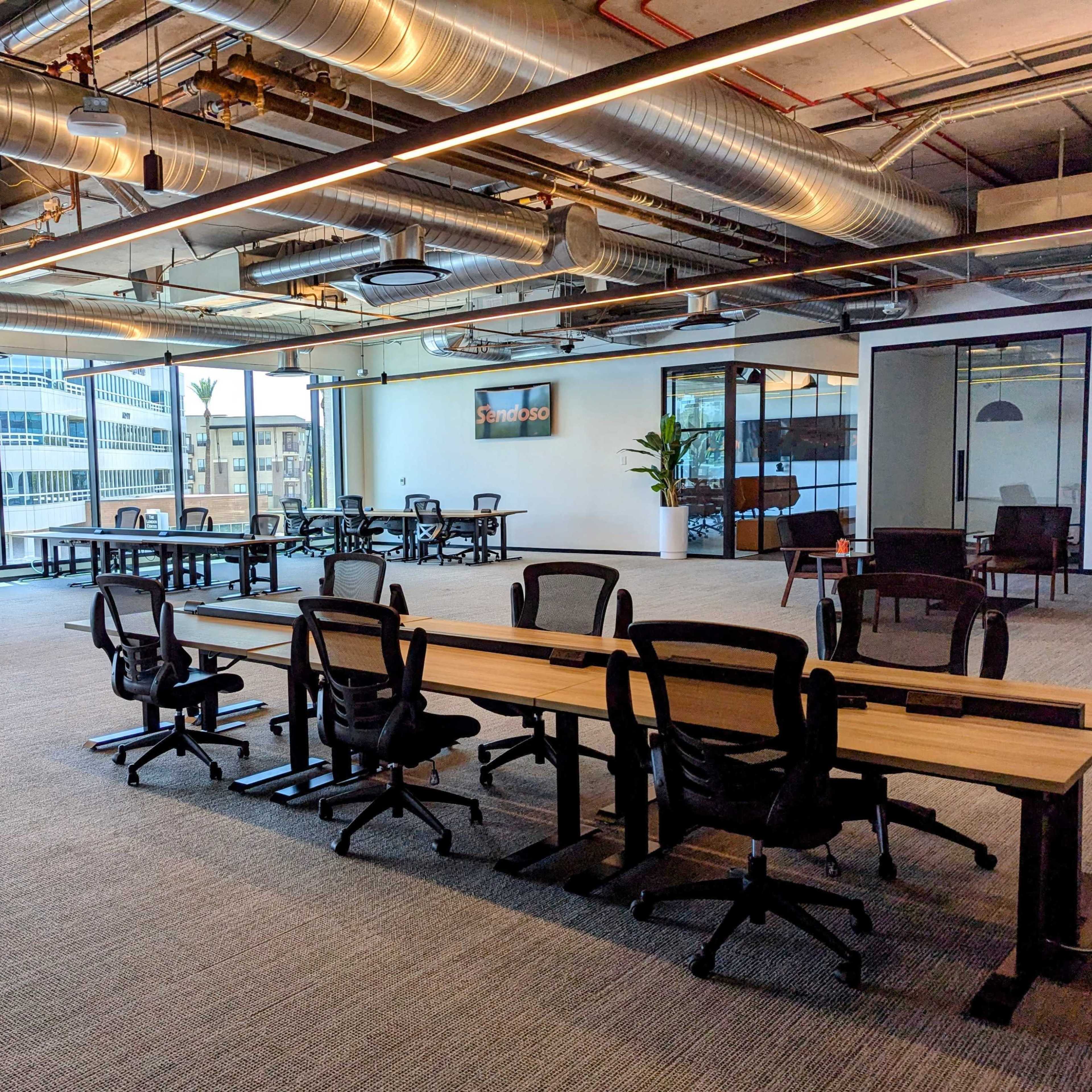 A modern office space with rows of empty desks, ergonomic chairs, and large windows allowing natural light.