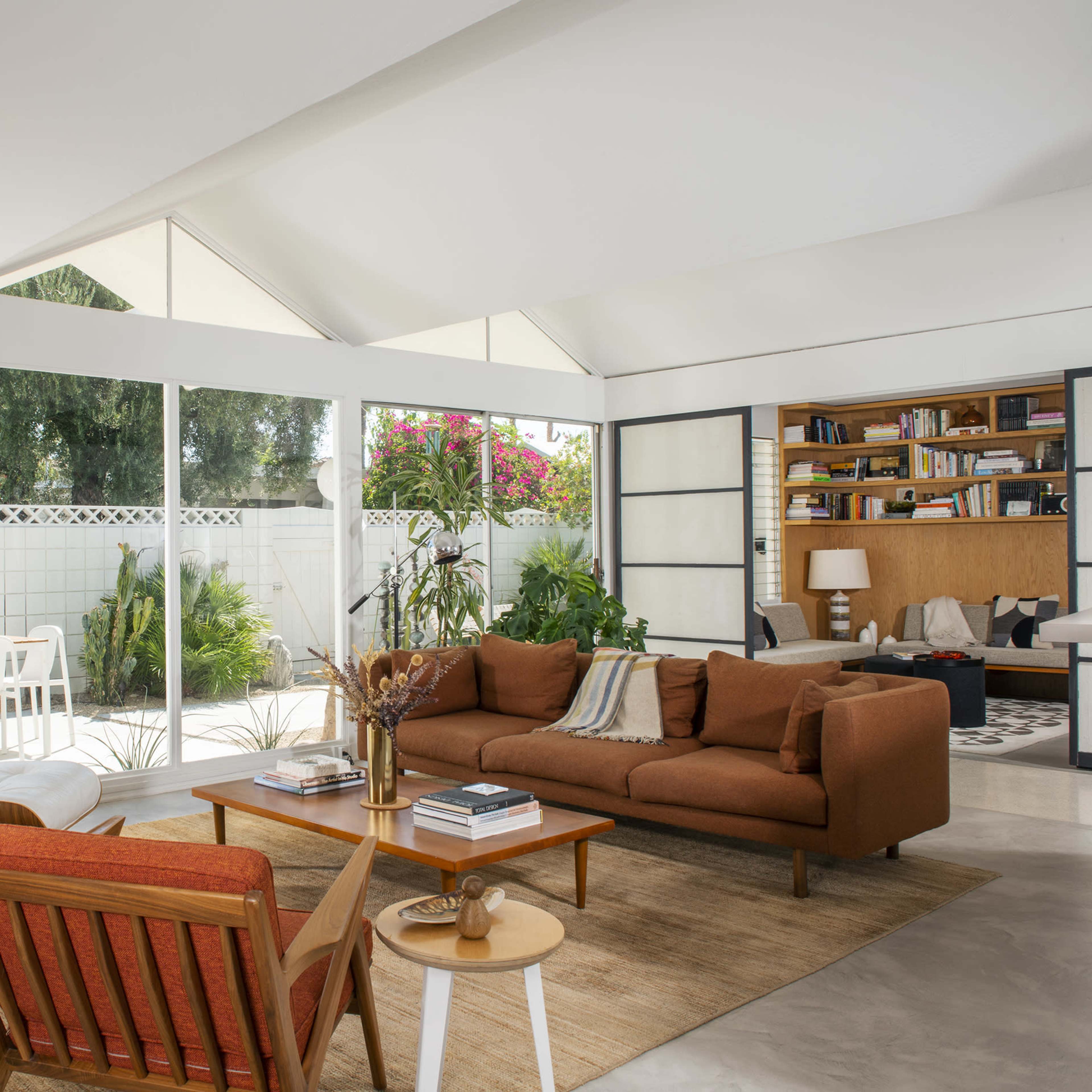 A modern living room features a brown sectional sofa, a pair of accent chairs, a coffee table, and large windows that provide a view of an outdoor patio and greenery.