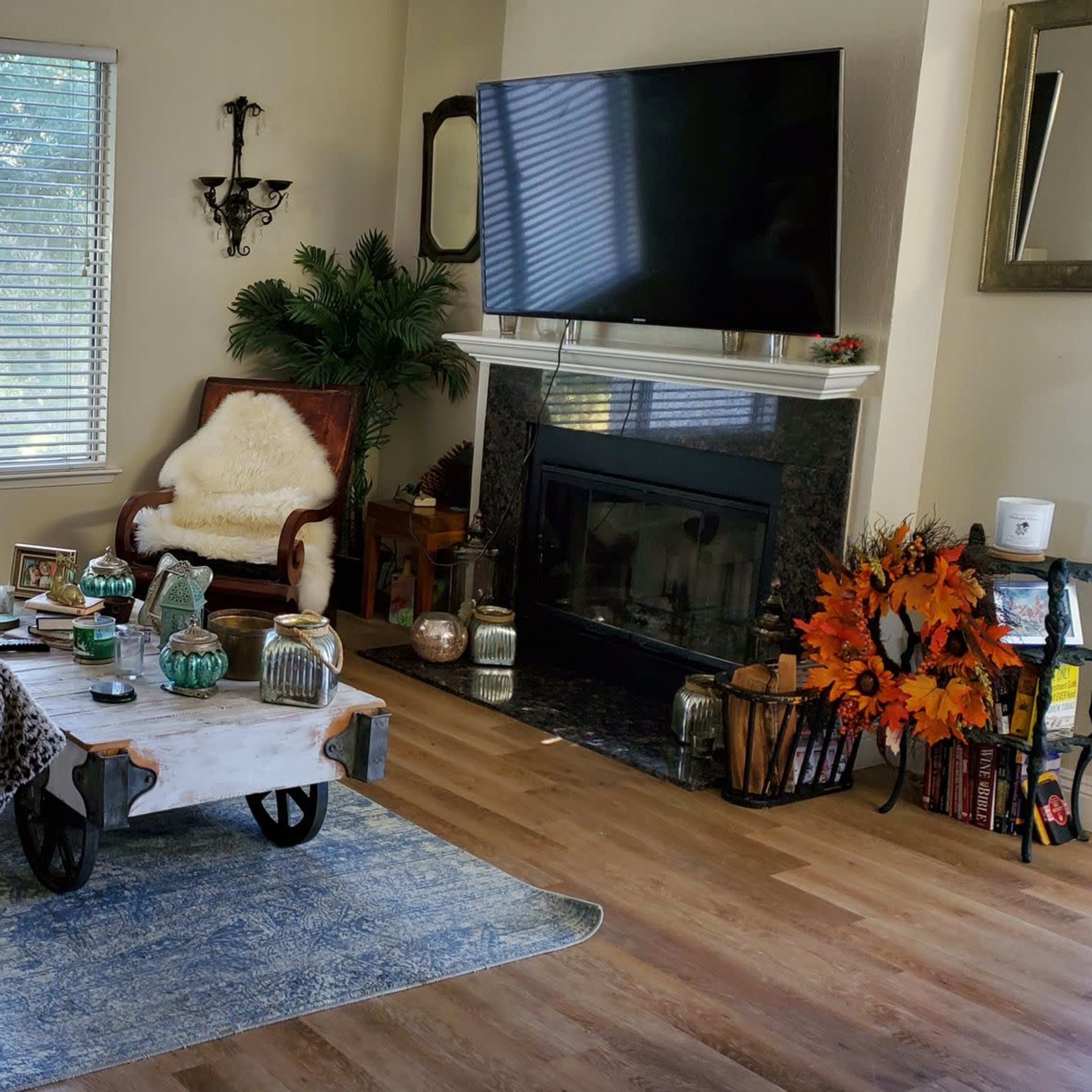 The living room features a cozy arrangement with a yellow sofa, a wooden coffee table, and a large television above a fireplace, all illuminated by natural light from a window.