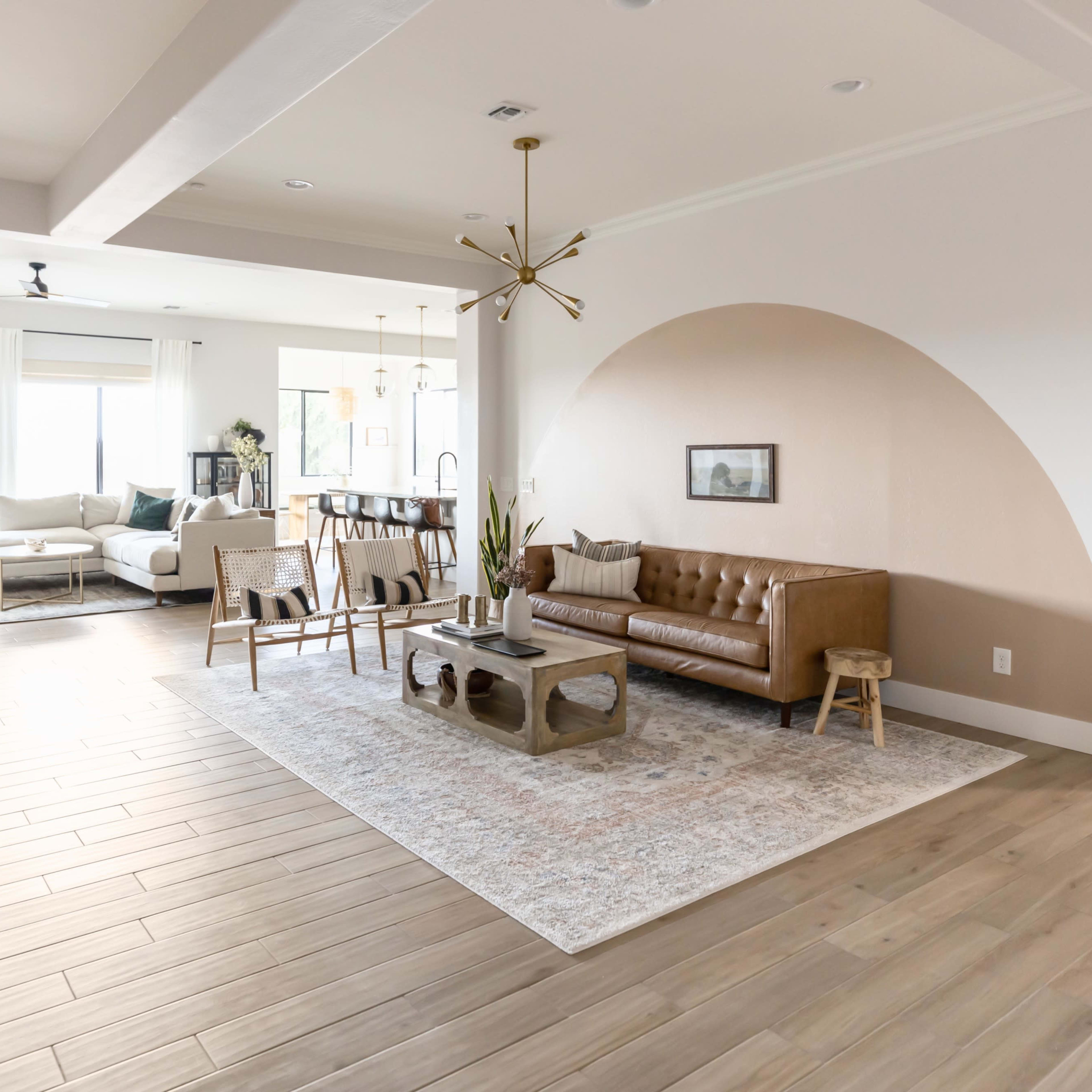 A spacious living room features a brown leather sofa, a rectangular coffee table, and large windows that allow natural light to fill the area.
