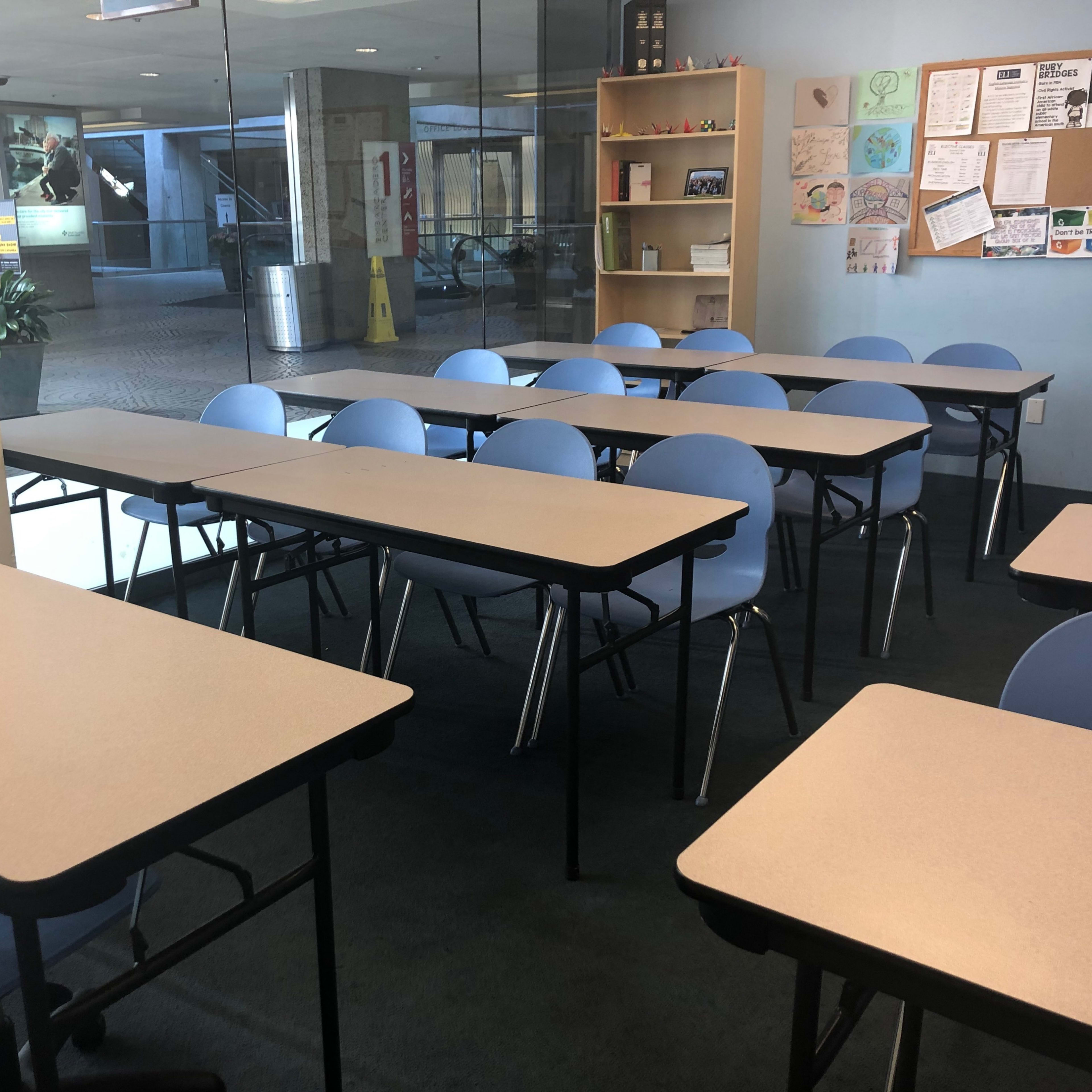 A classroom is set up with several rows of tables and blue chairs, illuminated by natural light coming through large windows.