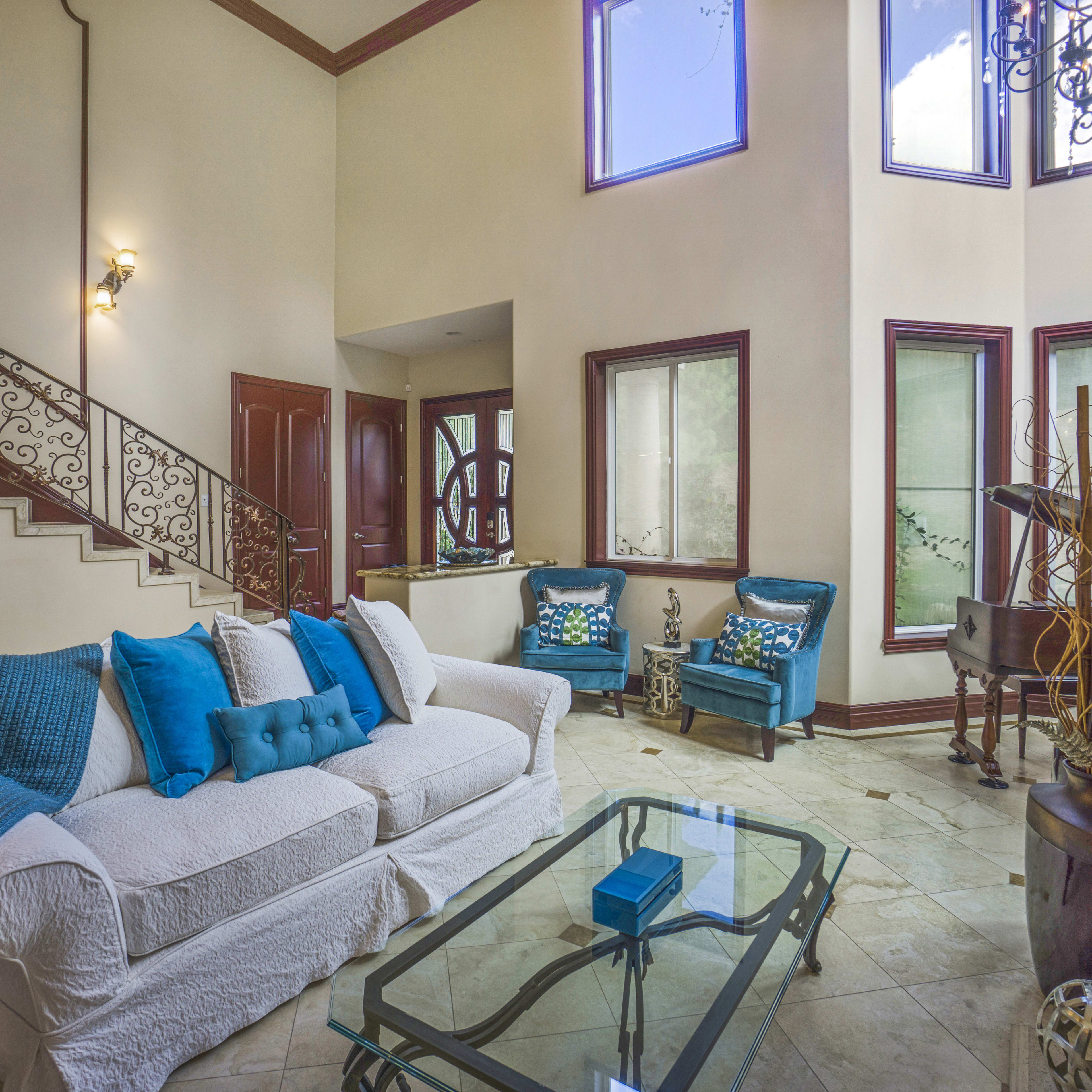 A well-lit living room featuring a white sofa with blue cushions, a glass coffee table, and large windows, along with a staircase and decorative elements.