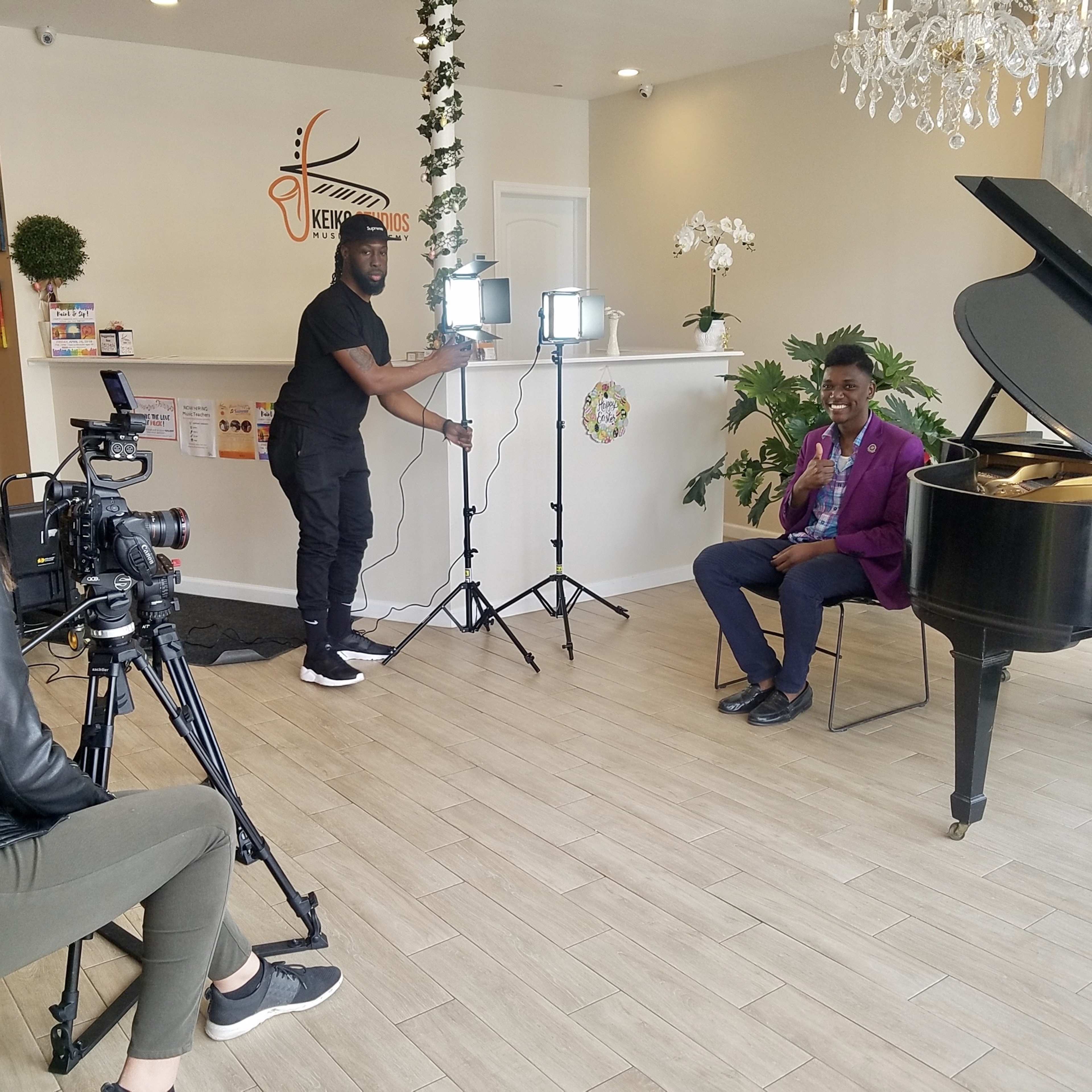 A man is seated at a grand piano while being filmed by a camera operator in a bright, indoor setting.