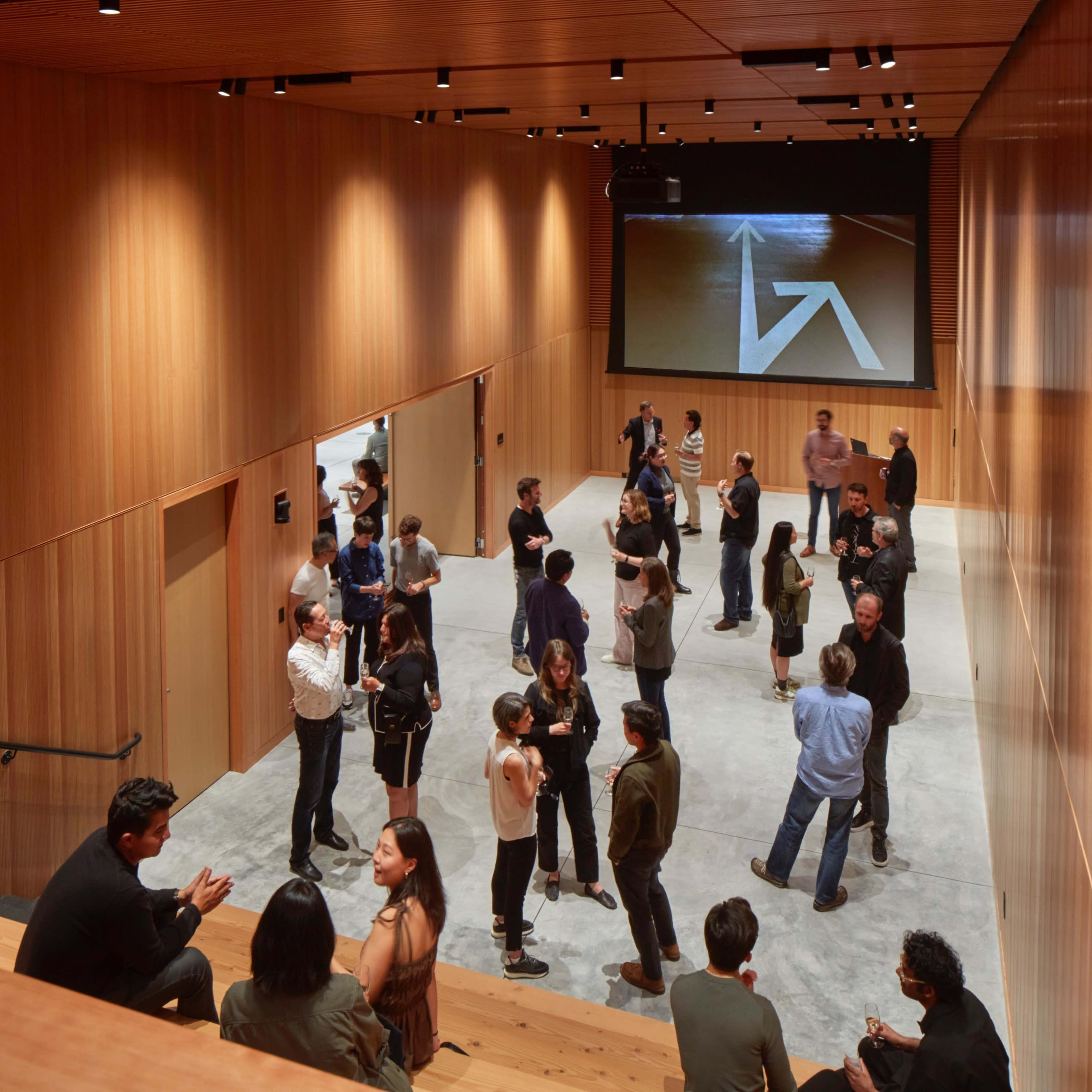 A group of people is gathered in a modern wooden interior space with a large screen displaying an arrow.