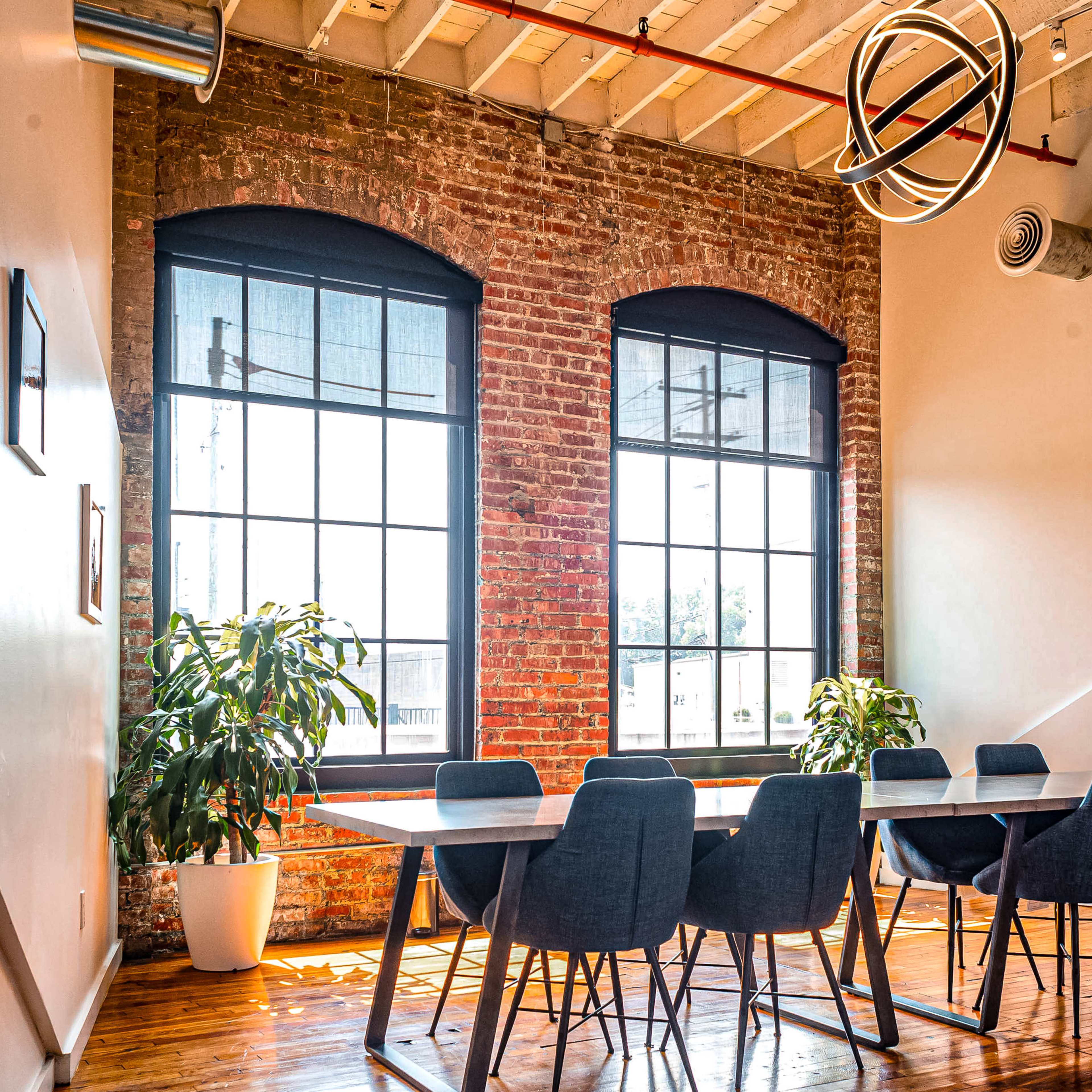 A bright meeting room features a long table surrounded by chairs, large windows with sheer curtains, and a potted plant in the corner.