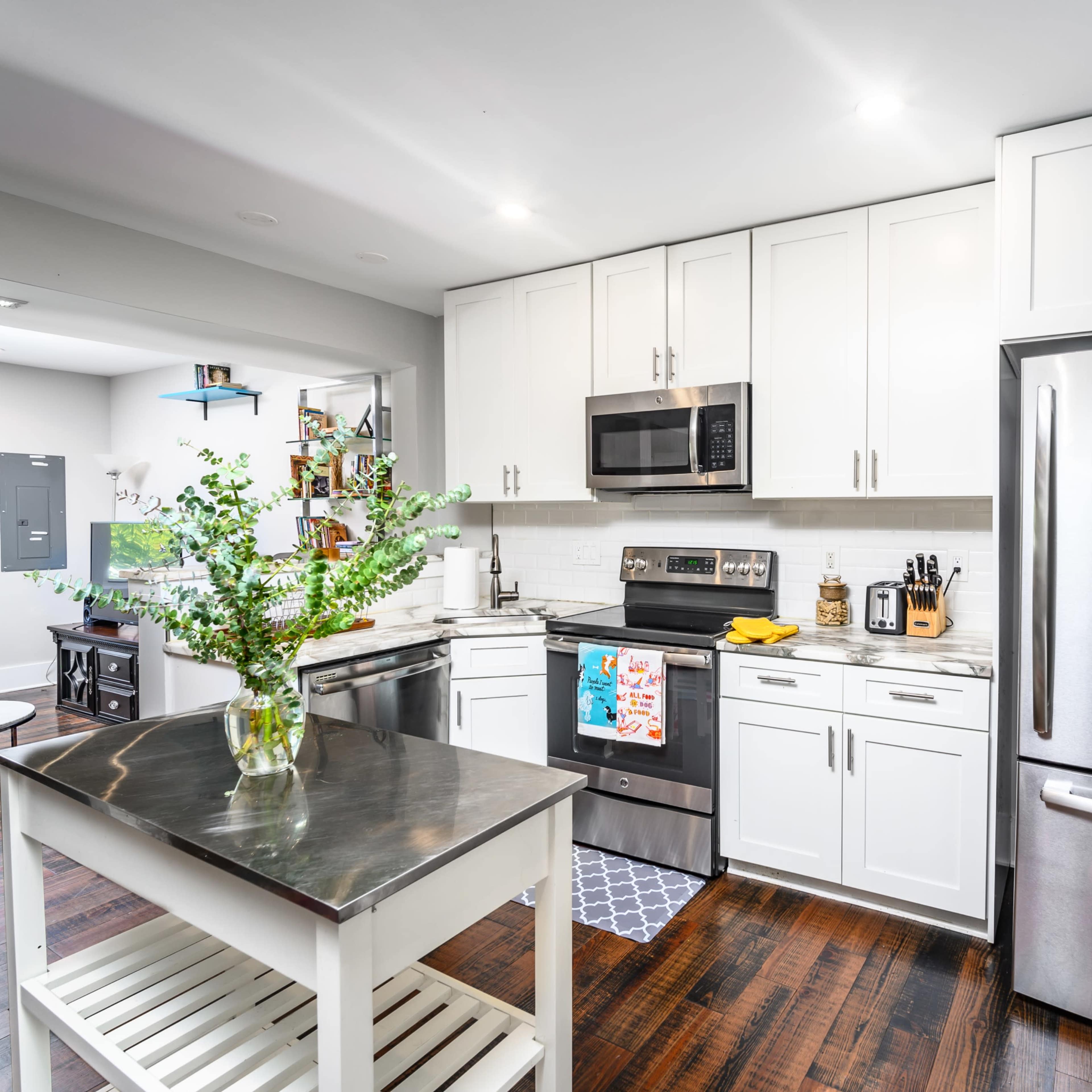 A modern kitchen features white cabinetry, stainless steel appliances, and a central island with a stainless steel top, complemented by wooden flooring.